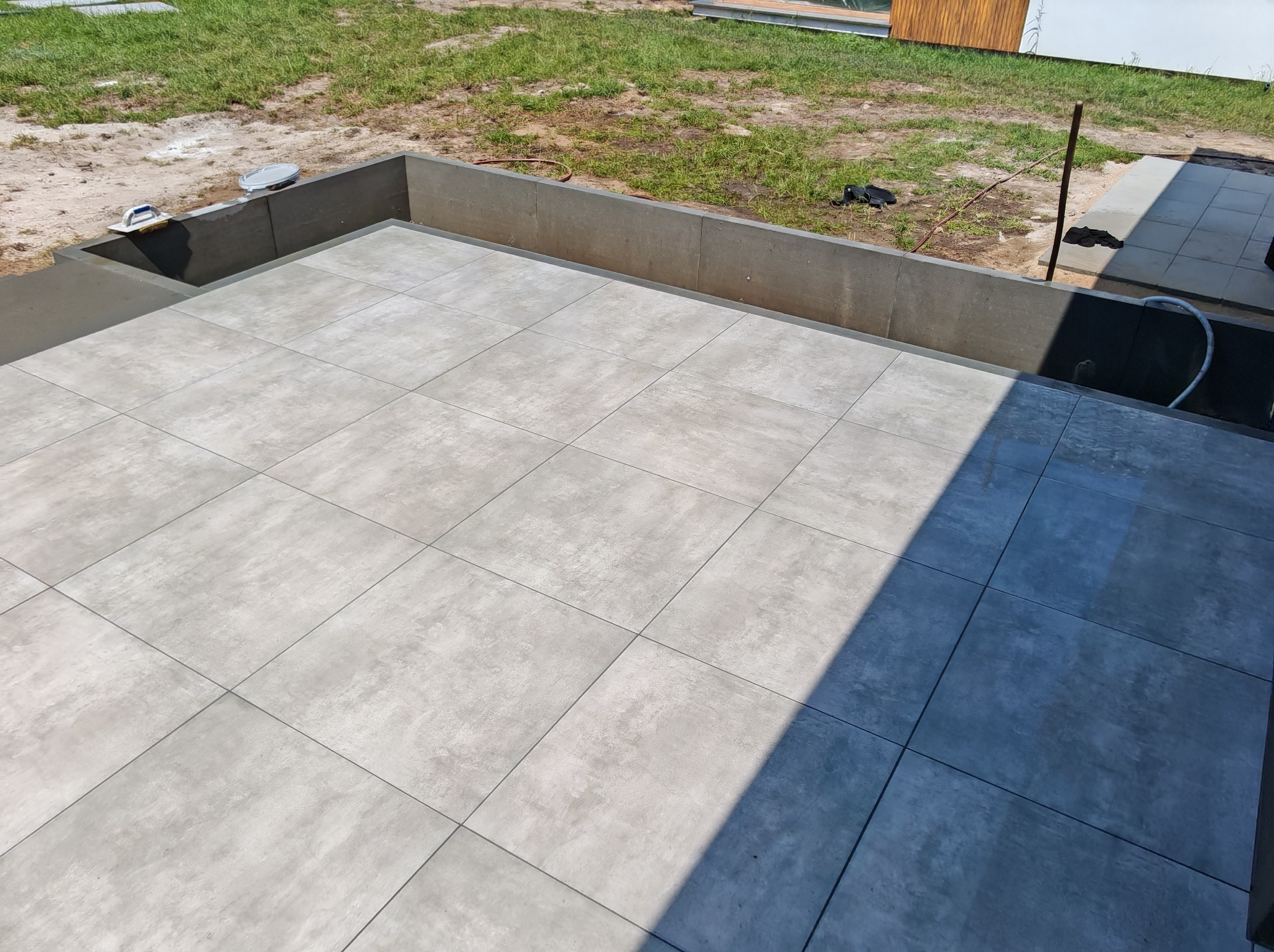 Ułożona, szara, kwadratowa płyta tarasowa z widocznymi narzędziami i fragmentem trawnika w tle. Płyty ułożone równo, z zachowaniem odstępów.