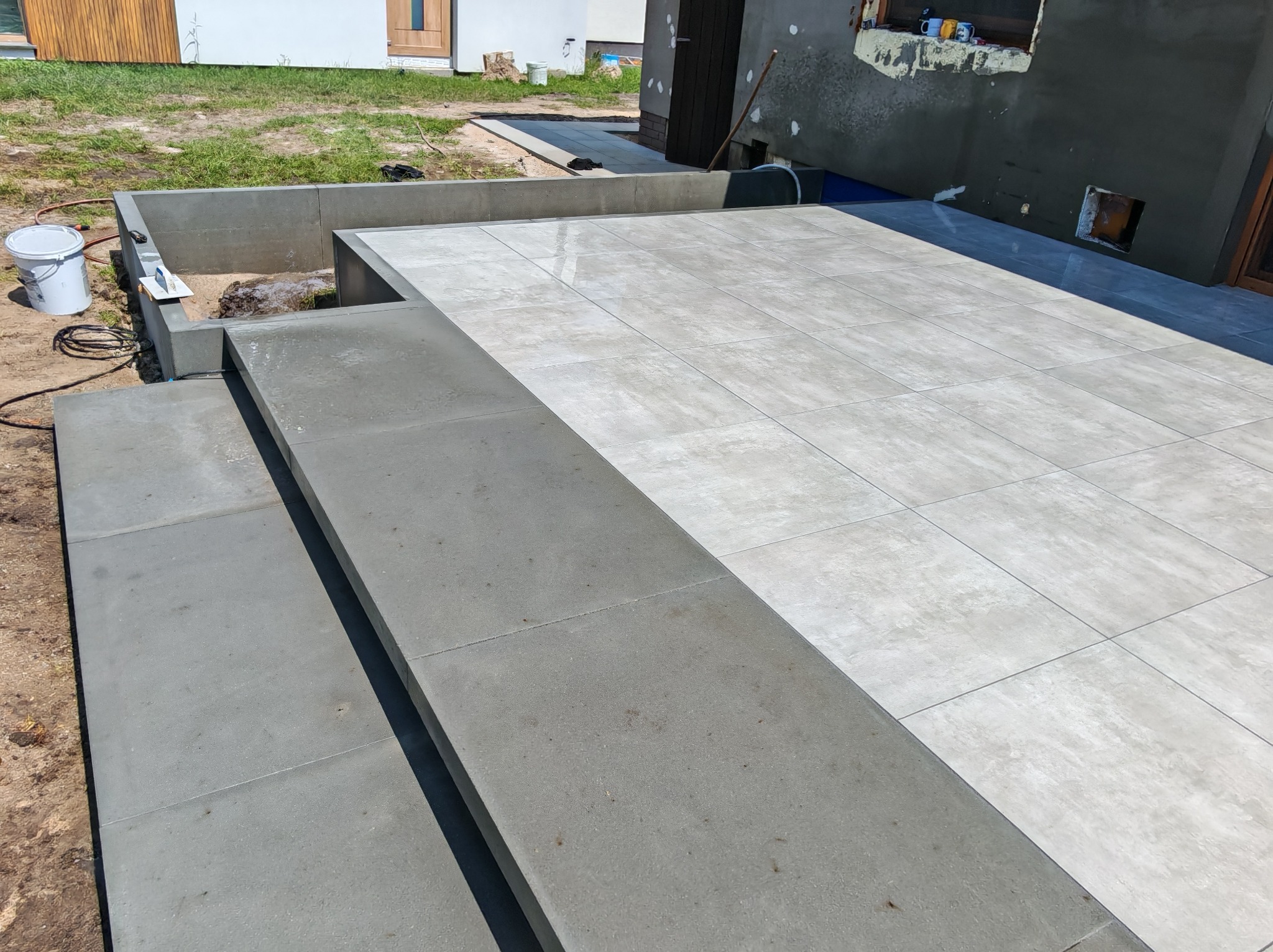 Wykończony taras z jasnych, kwadratowych płyt. Widoczne betonowe obrzeża i schody. W tle trawnik i fragment budynku w trakcie prac wykończeniowych.