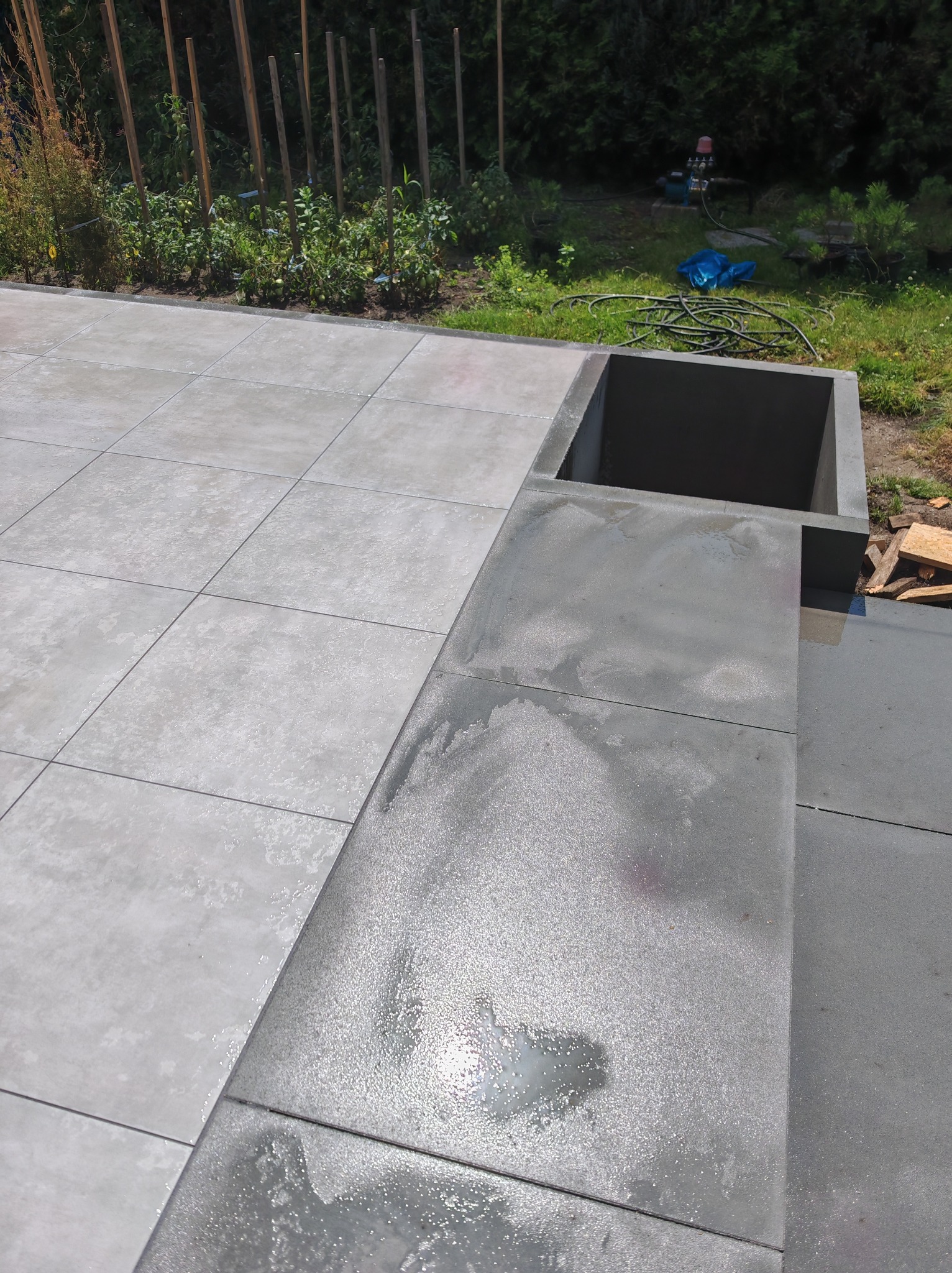 Nowoczesny taras z szarych płyt betonowych, z fragmentem rabaty i elementem małej architektury z betonu. Widoczny fragment ogrodu z roślinnością.