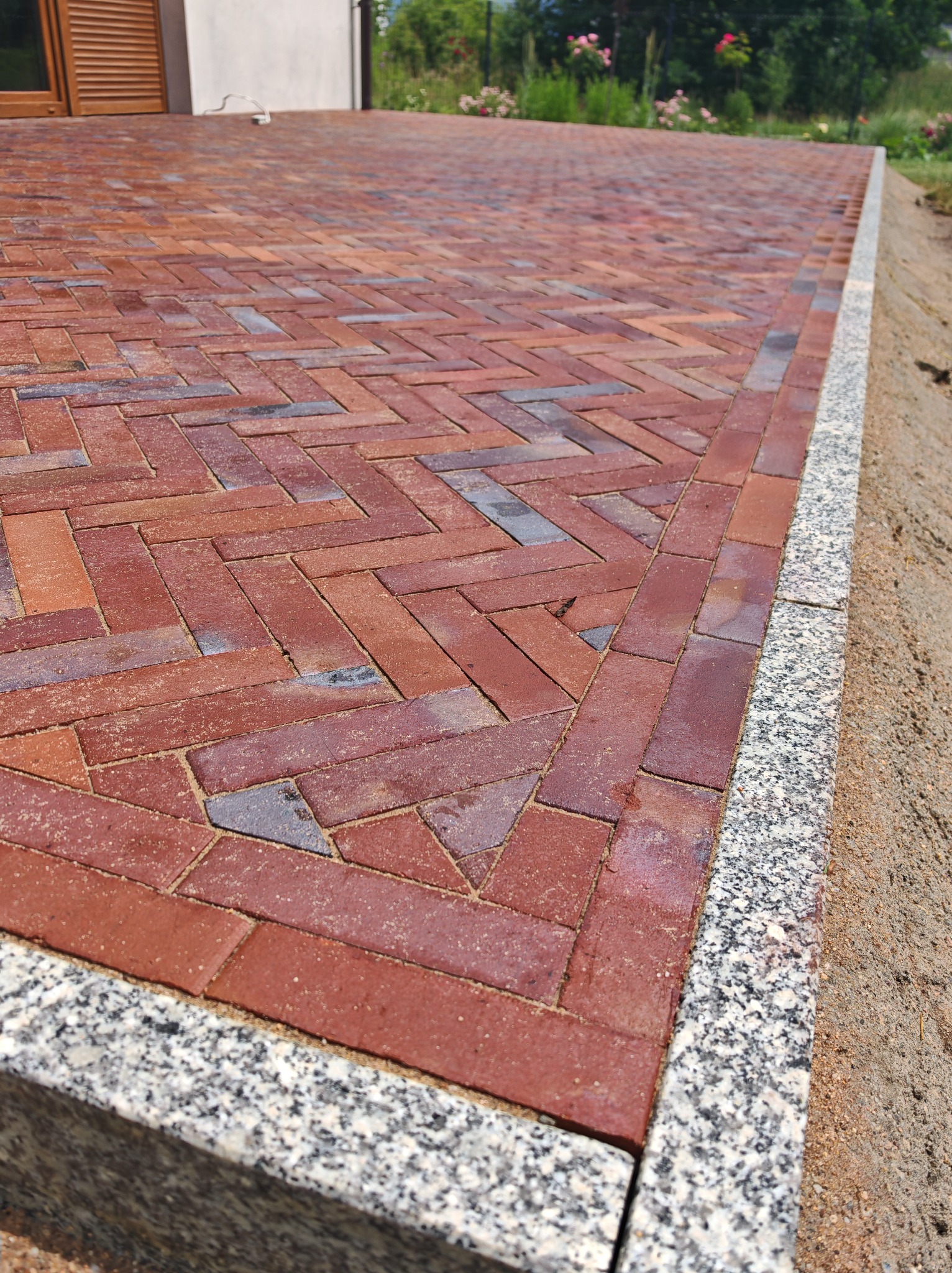 Ceglana kostka brukowa ułożona w jodełkę, obrzeża z jasnego granitu. Widok z góry, lekko z boku, ukazujący wzór i detale wykończenia.