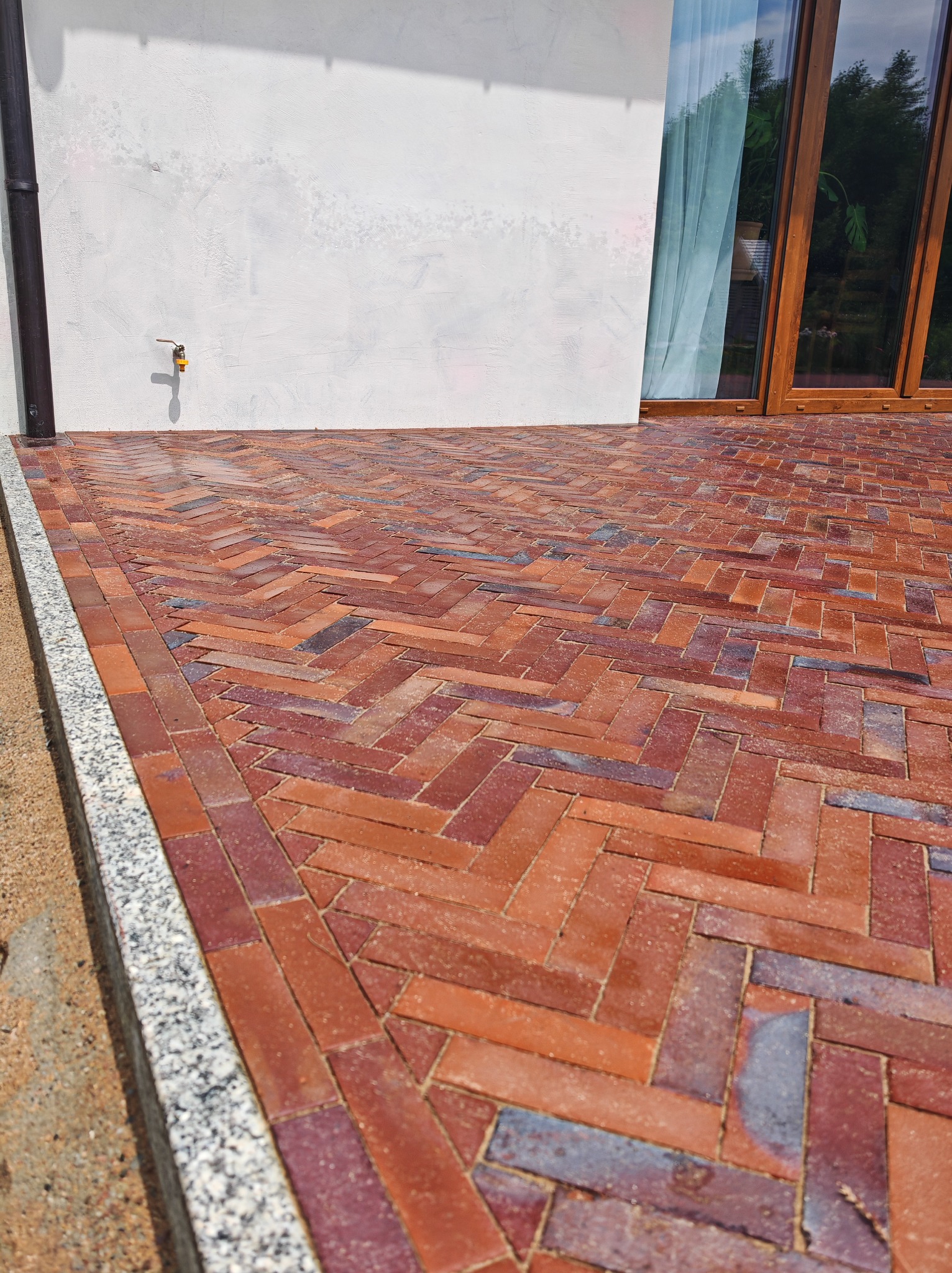 Ceglana kostka brukowa ułożona w jodełkę na posesji, obrzeże z jasnego granitu, widoczna ściana budynku i okno balkonowe. Detal wykończenia nawierzchni.