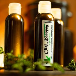 TOVPIKART Robert Towpik - Trzy butelki oleju konopnego z żywokostem na drewnianym blacie, otoczone suszem konopnym, w ciepłym oświetleniu. Etykieta z zielonym akcentem i napisem 'Olej Konopny'.