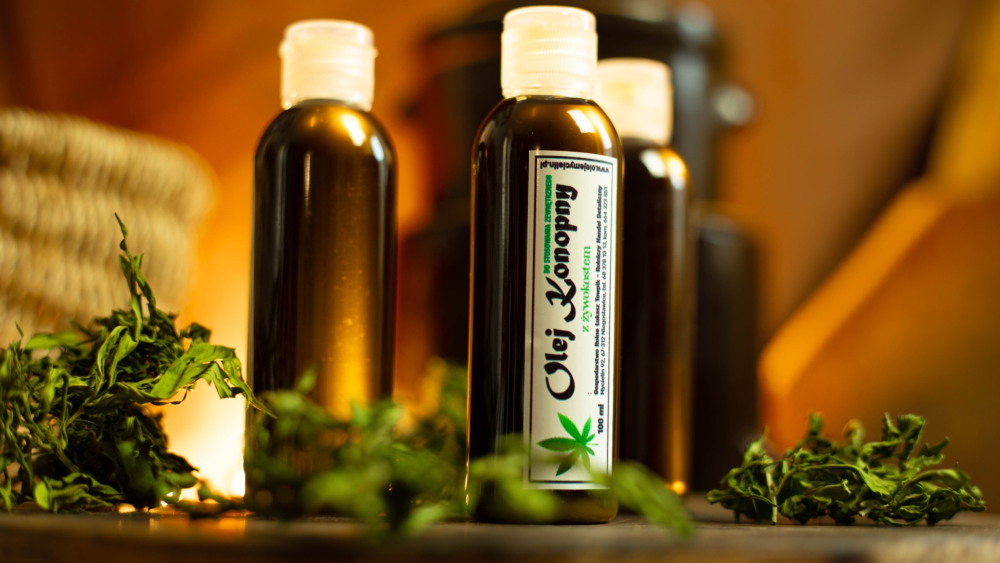 Trzy butelki oleju konopnego z żywokostem na drewnianym blacie, otoczone suszem konopnym, w ciepłym oświetleniu. Etykieta z zielonym akcentem i napisem 'Olej Konopny'.