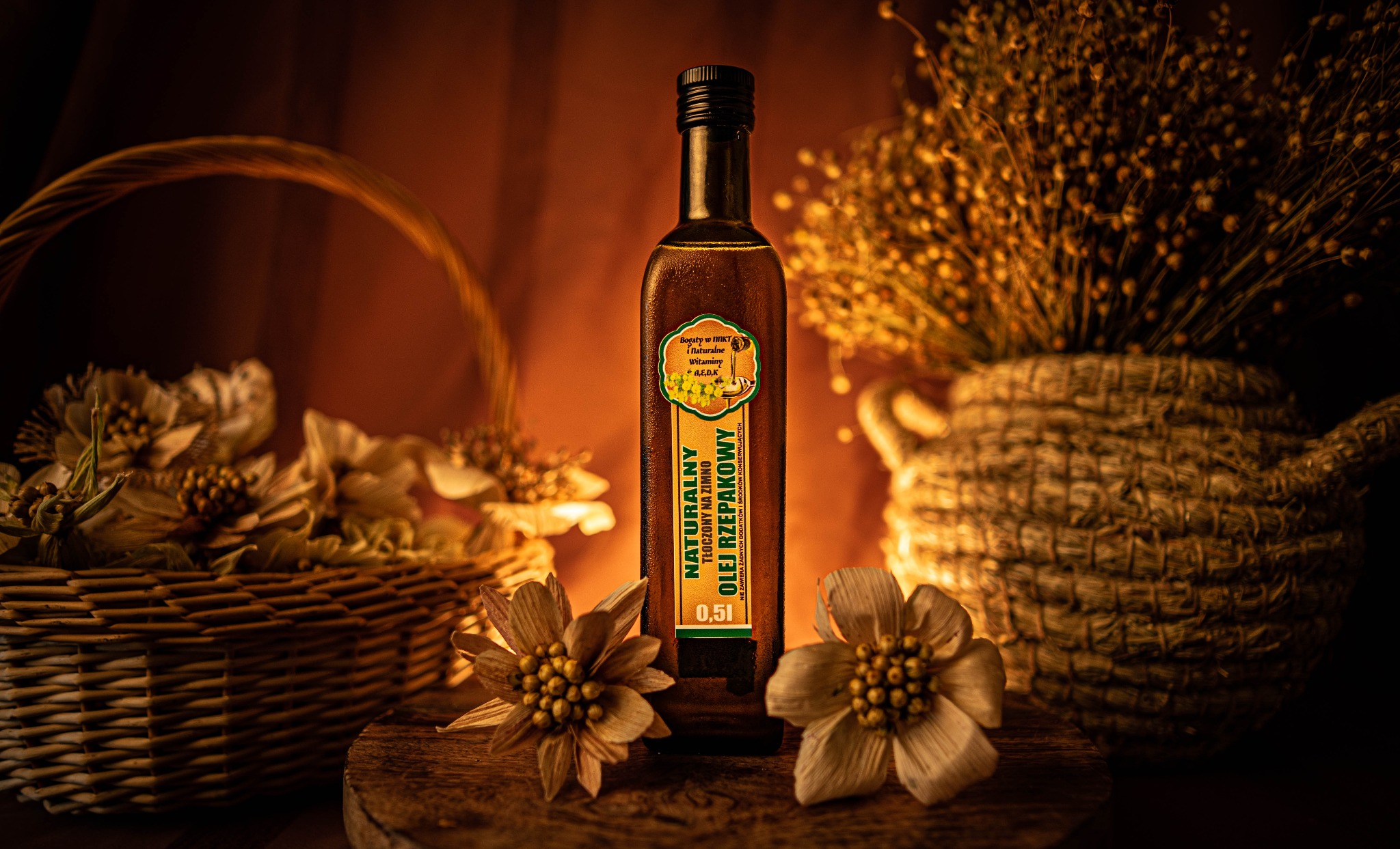Butelka oleju rzepakowego 0.5l na drewnianej desce, otoczona kwiatami i koszykami w ciepłym oświetleniu. Produkt spożywczy w aranżacji.