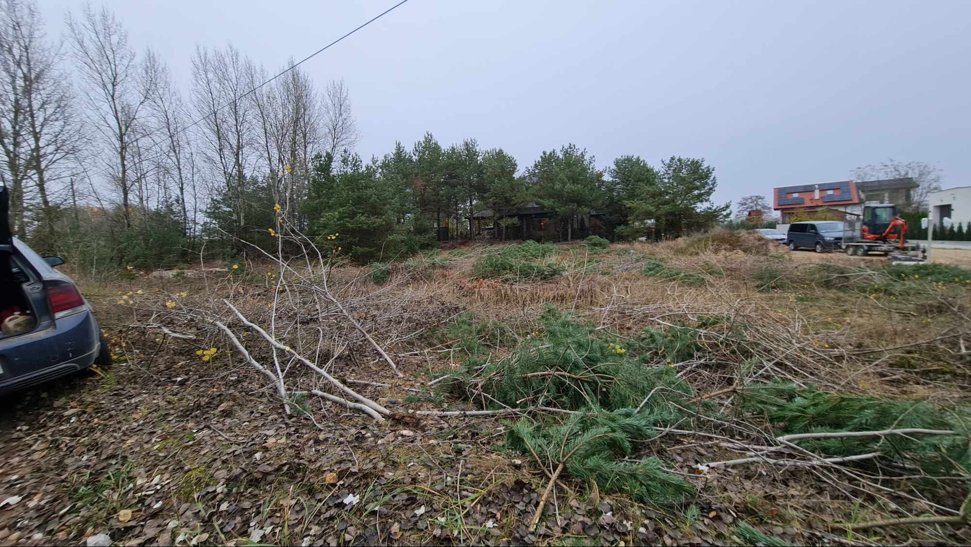 Teren po wycince drzew i krzewów, widoczne gałęzie na ziemi, w tle budynek z panelami słonecznymi i sprzęt budowlany.