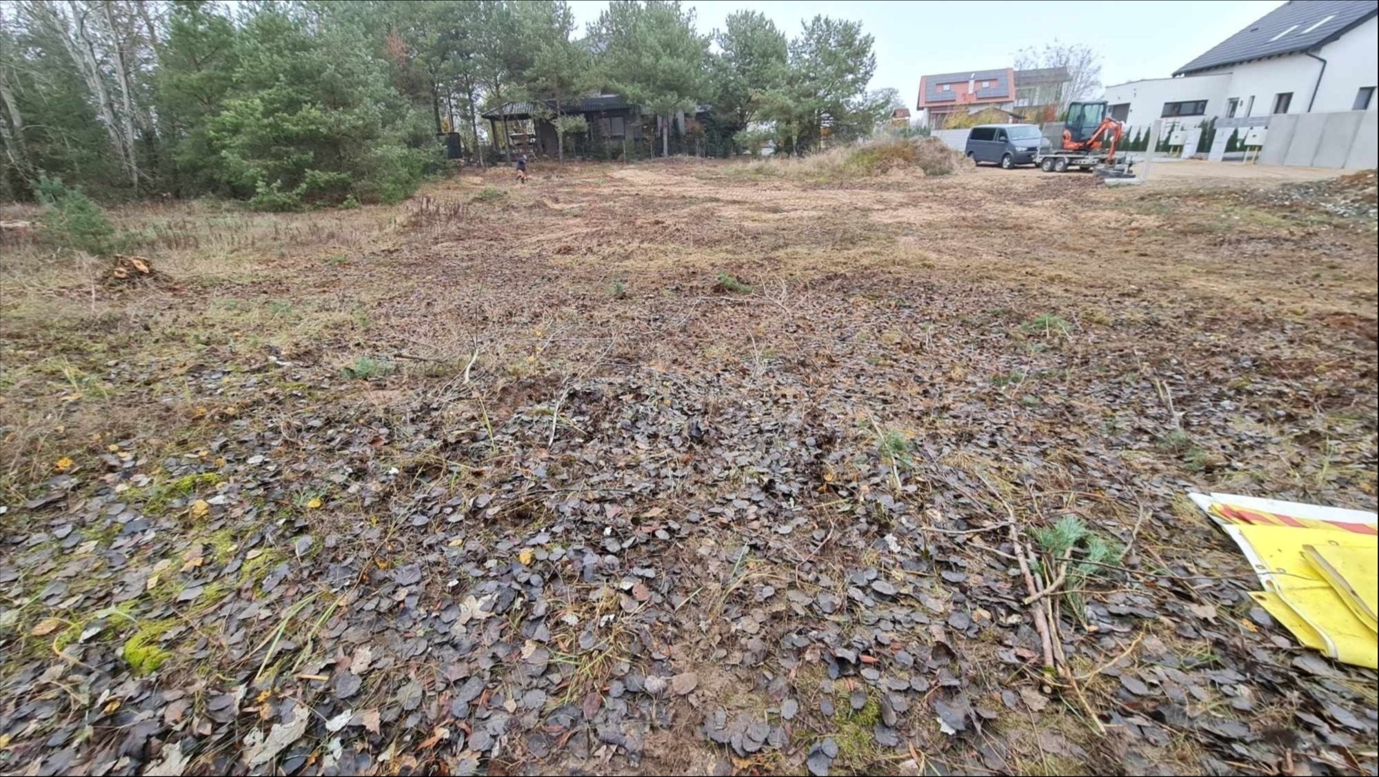 Teren porośnięty suchą trawą i liśćmi, z widocznymi drzewami w tle oraz mini koparką i samochodem, prawdopodobnie przygotowywany pod inwestycję.