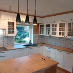 Nowoczesna kuchnia z białymi szafkami z przeszkleniami, drewnianymi blatami i wyspą, oświetlona trzema wiszącymi lampami z czarnymi kloszami.