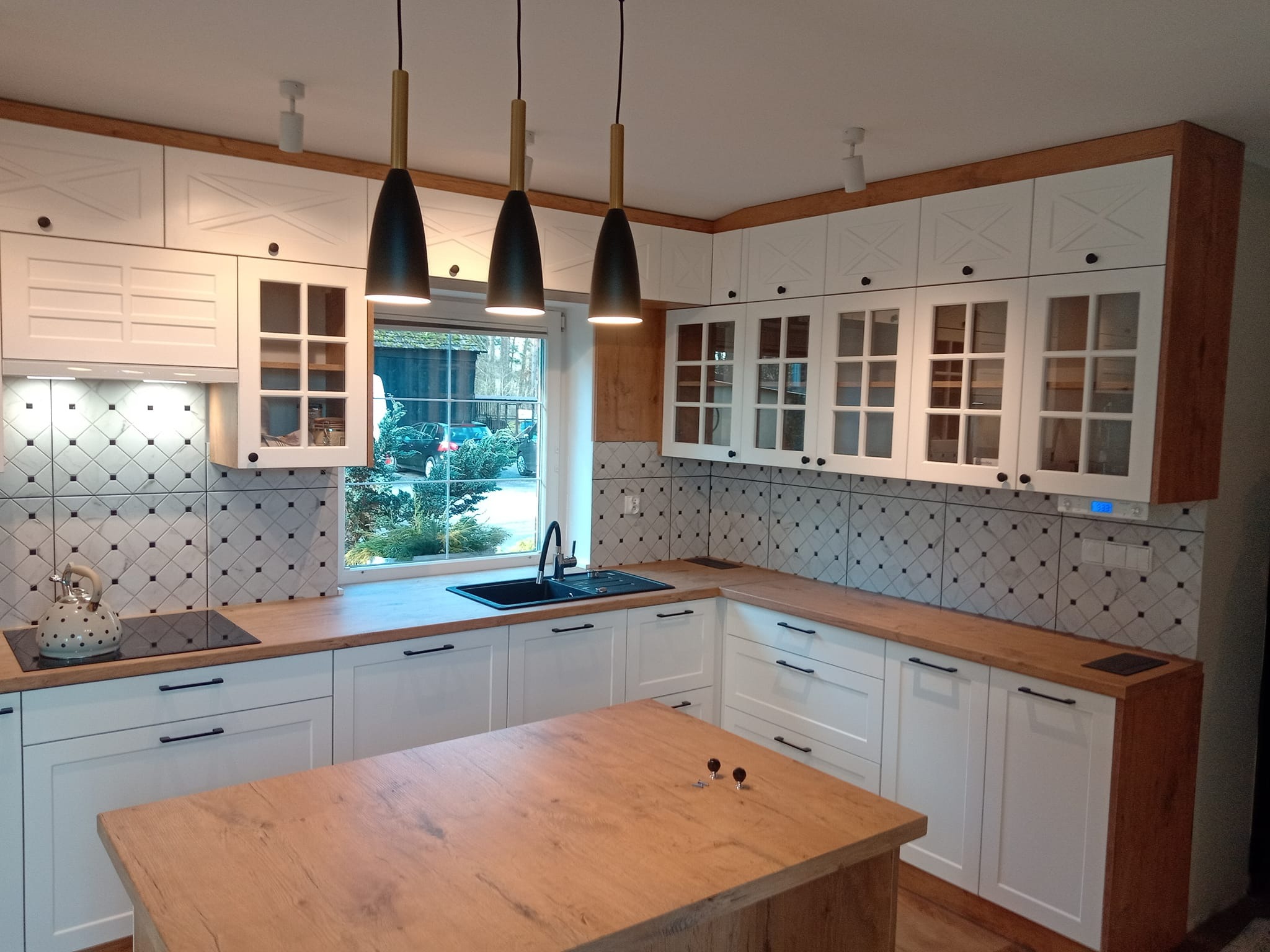 Nowoczesna kuchnia z białymi szafkami z przeszkleniami, drewnianymi blatami i wyspą, oświetlona trzema wiszącymi lampami z czarnymi kloszami.