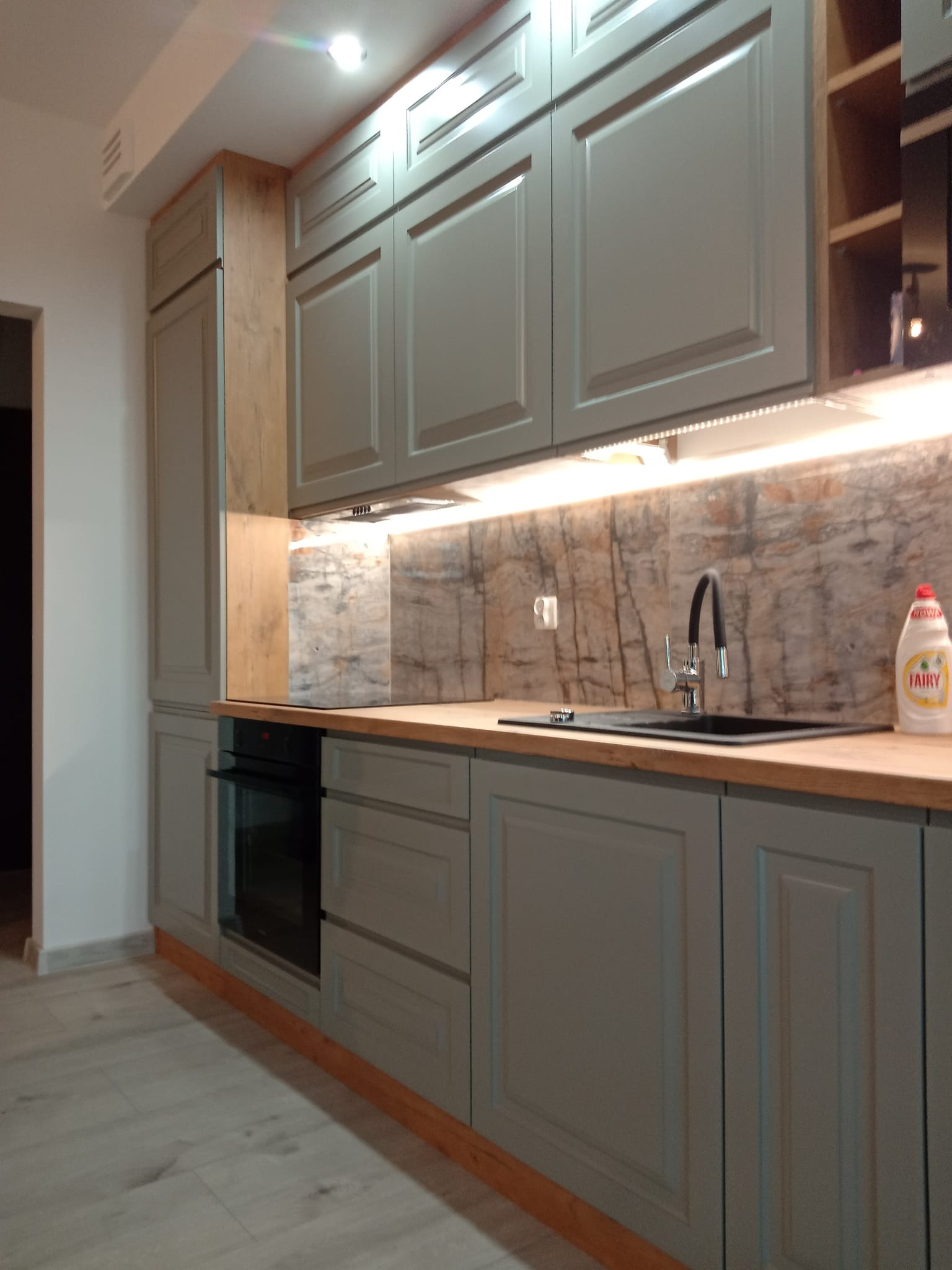 Szare meble kuchenne z drewnianym blatem i marmurowym panelem nad zlewem, oświetlenie LED pod szafkami wiszącymi, widoczna butelka płynu do naczyń.