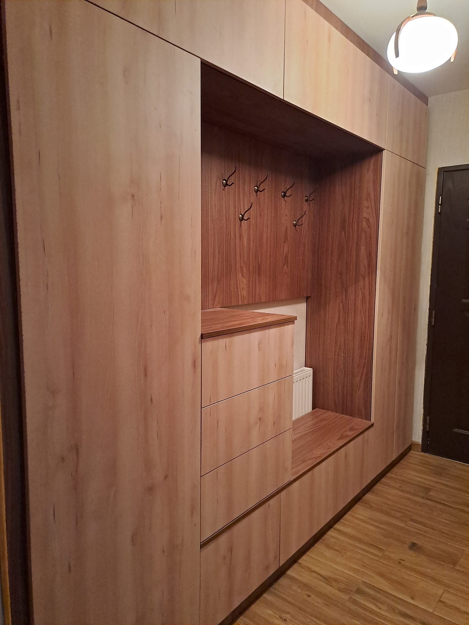 Zabudowa przedpokoju z jasnego drewna z wnęką na siedzisko i wieszakami, widoczne trzy szuflady i szafki górne.