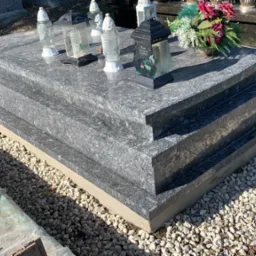 Granitowy pomnik z trzema poziomami, ozdobiony lampionami i kwiatami, otoczony jasnym żwirem.