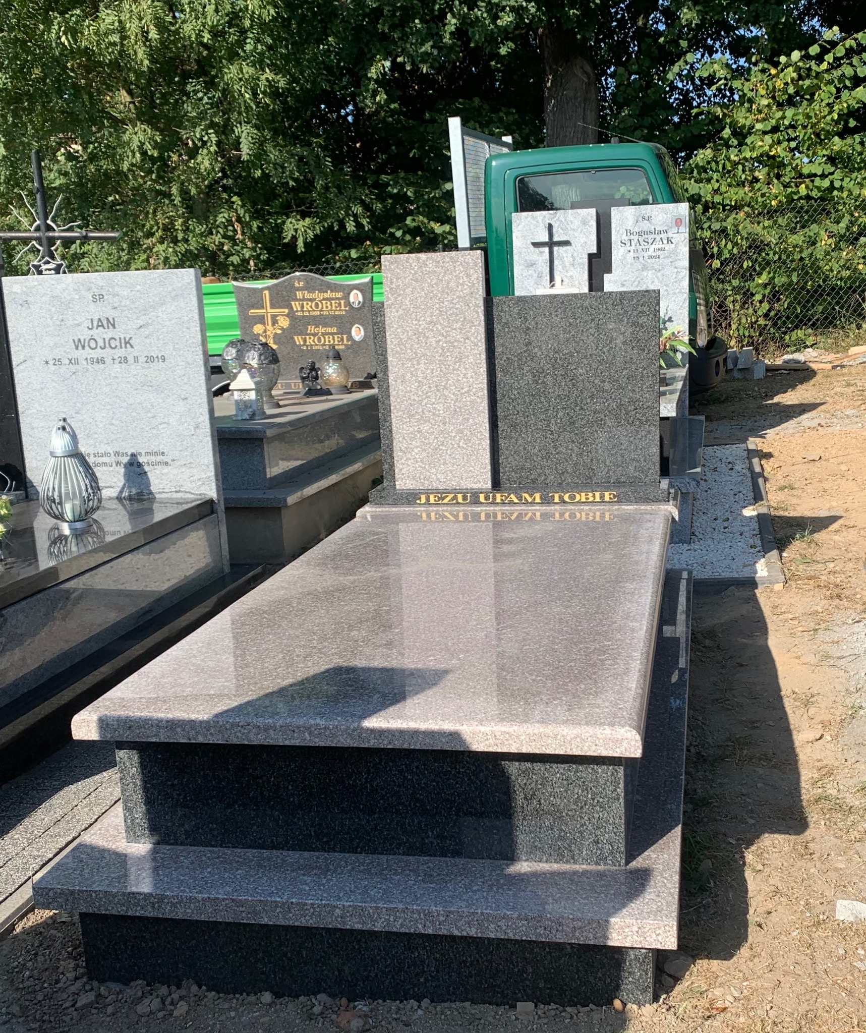 Nowoczesny, trzyczęściowy pomnik z jasnego i ciemnego granitu z napisem 'Jezu Ufam Tobie' w złotych literach, ustawiony na cmentarzu w słoneczny dzień, z widocznym zielonym samochodem w tle.