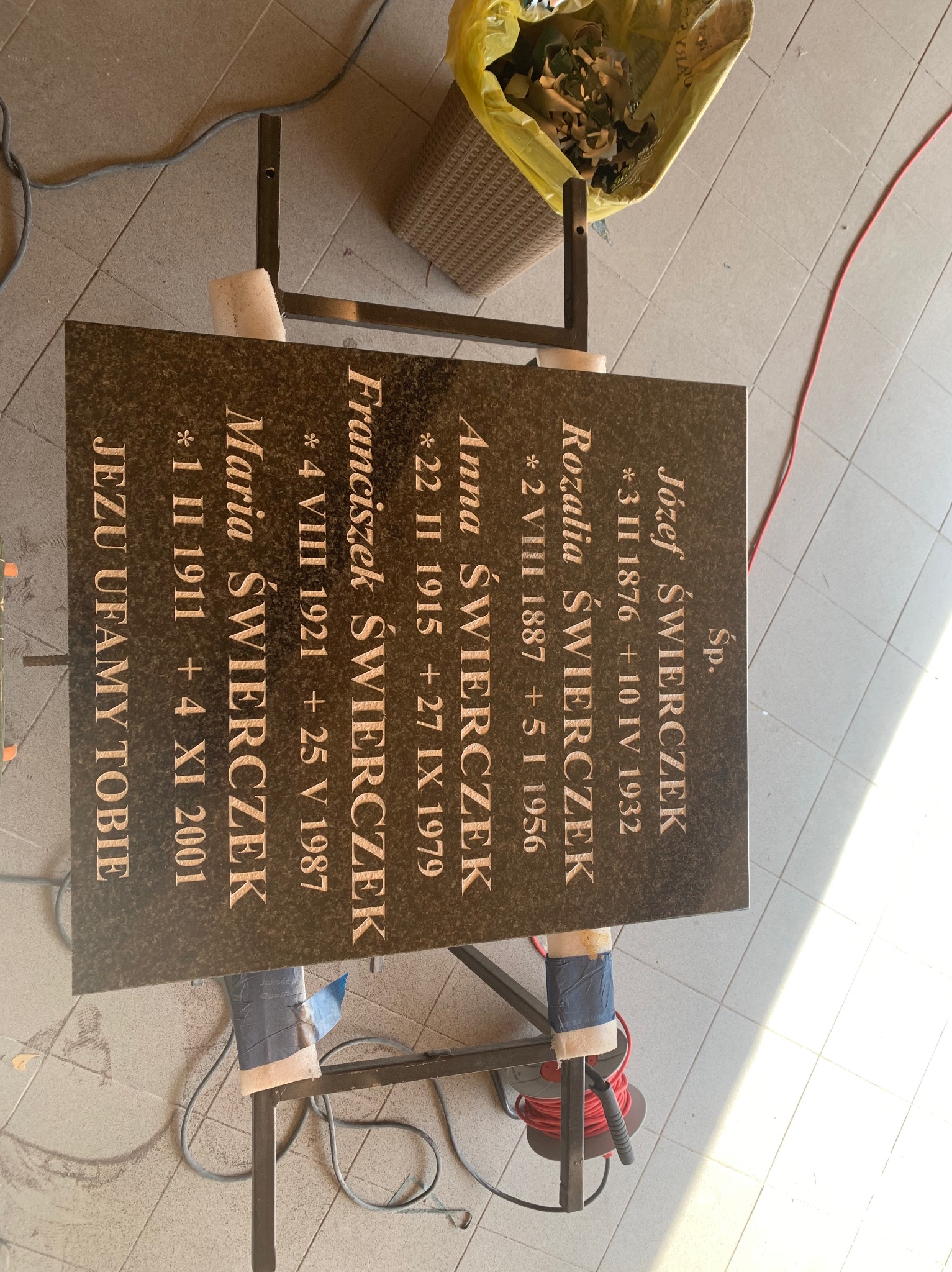 Brązowa płyta nagrobna z wygrawerowanymi złotymi literami, zawierająca imiona, nazwiska i daty urodzin oraz śmierci członków rodziny Świerczek, ustawiona na metalowym stojaku w warsztacie...