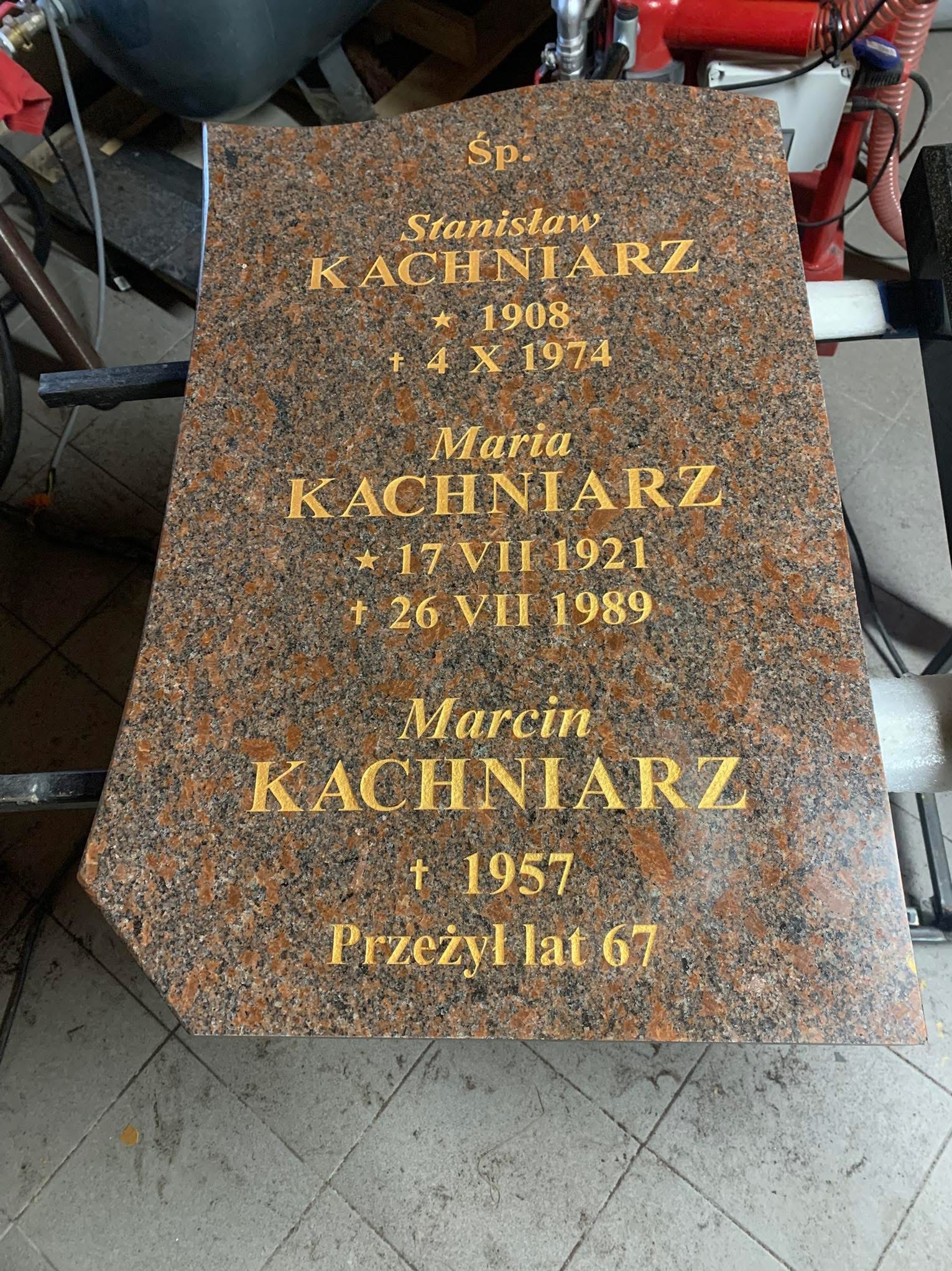 Brązowa płyta nagrobna z wygrawerowanymi złotymi literami, zawierająca imiona, nazwiska i daty zmarłych, ustawiona na metalowym stojaku w warsztacie kamieniarskim.