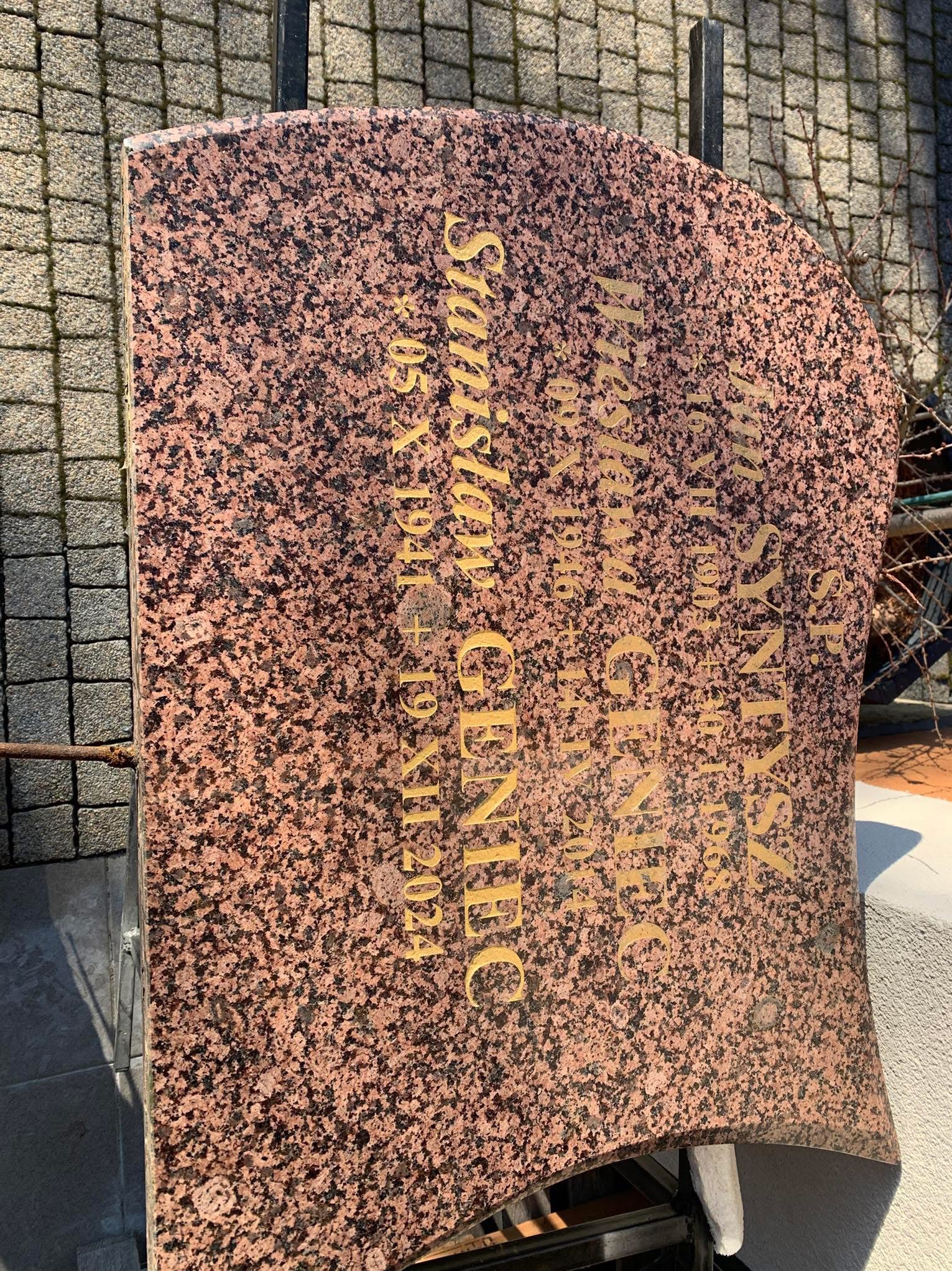 Granitowy nagrobek z wygrawerowanymi złotymi literami, widoczny fragment otoczenia cmentarza.