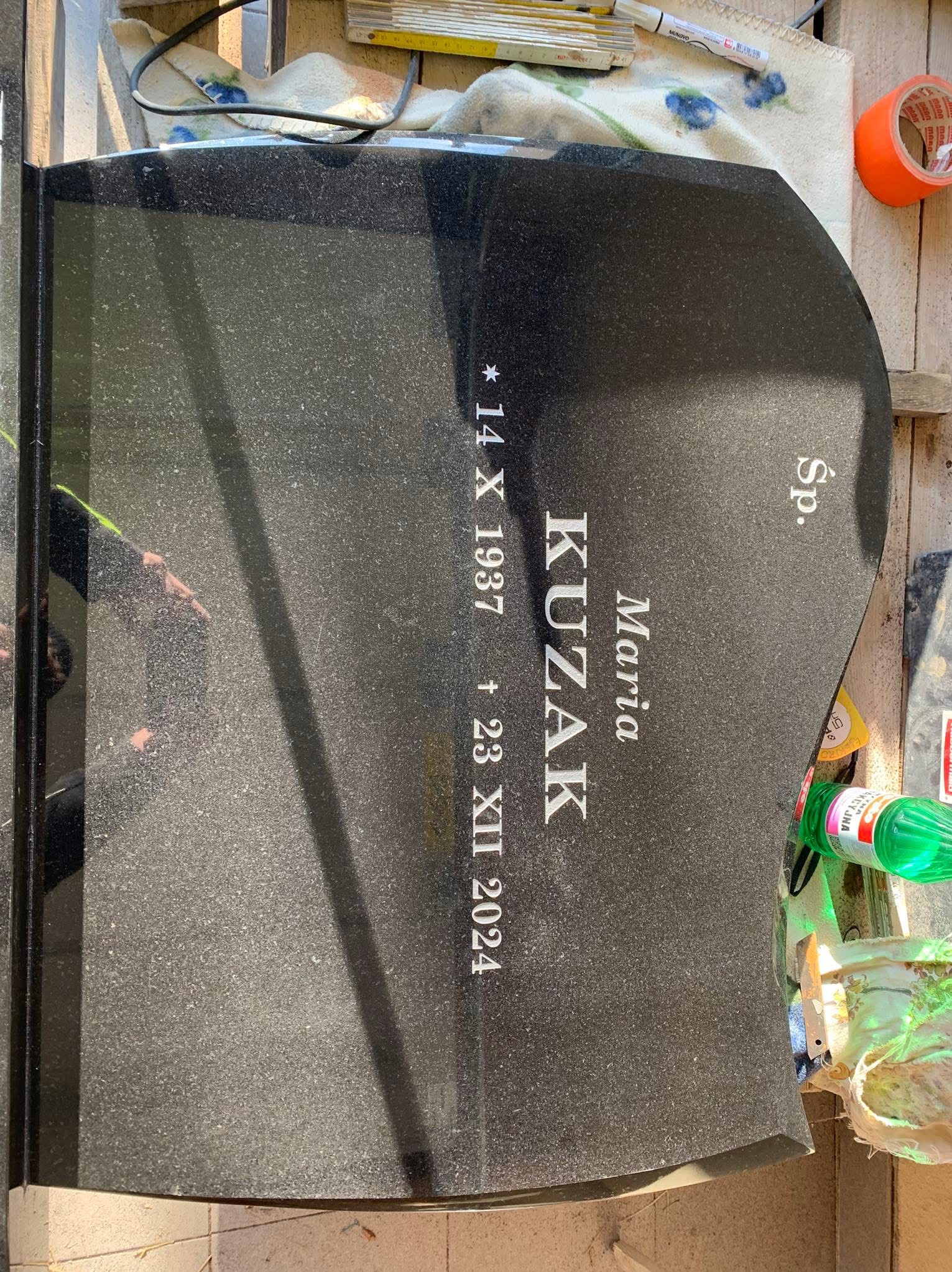 Czarny, granitowy pomnik z wygrawerowanym imieniem i nazwiskiem Maria Kuzak oraz datami urodzenia i śmierci, leżący na powierzchni roboczej z narzędziami i materiałami.