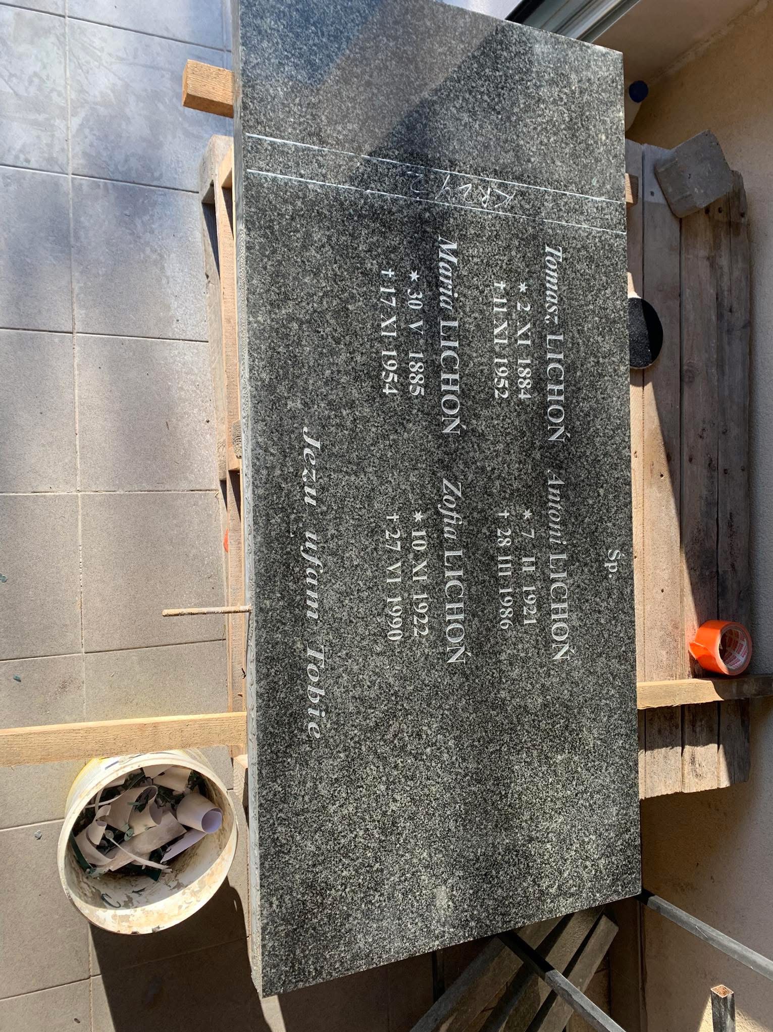 Granitowa płyta nagrobna z wygrawerowanymi danymi zmarłych, oparta o drewniane elementy podczas prac kamieniarskich, widoczny kubeł z odpadami po obróbce kamienia i rolka taśmy.