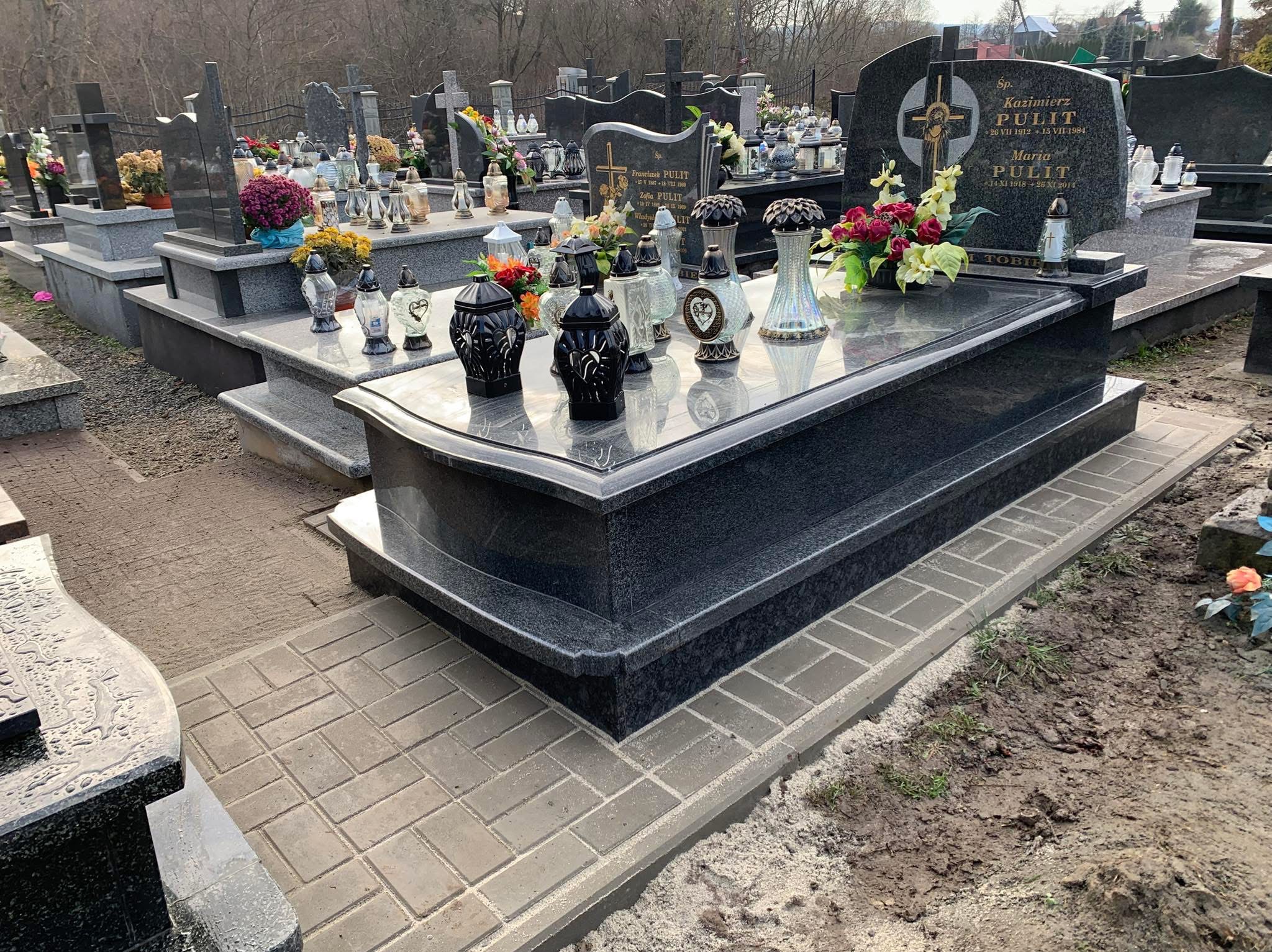 Ciemny, wypolerowany nagrobek z czarnego granitu, z licznymi lampionami i kwiatami, otoczony brukowaną ścieżką na cmentarzu, w tle inne nagrobki i drzewa.