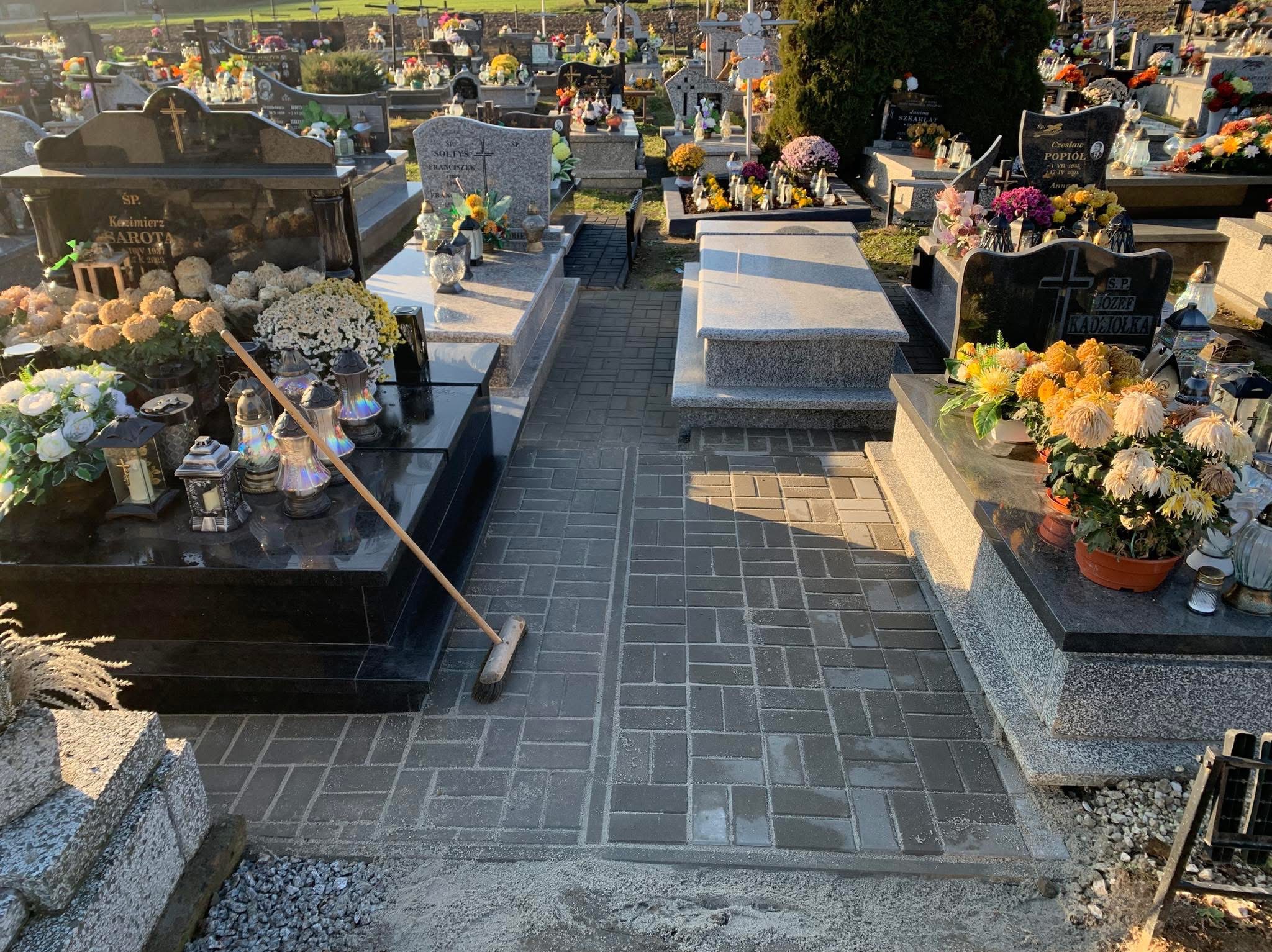 Częściowo ułożona szara kostka brukowa między nagrobkami na cmentarzu, widoczna miotła oparta o jeden z pomników, w tle liczne groby i zieleń.