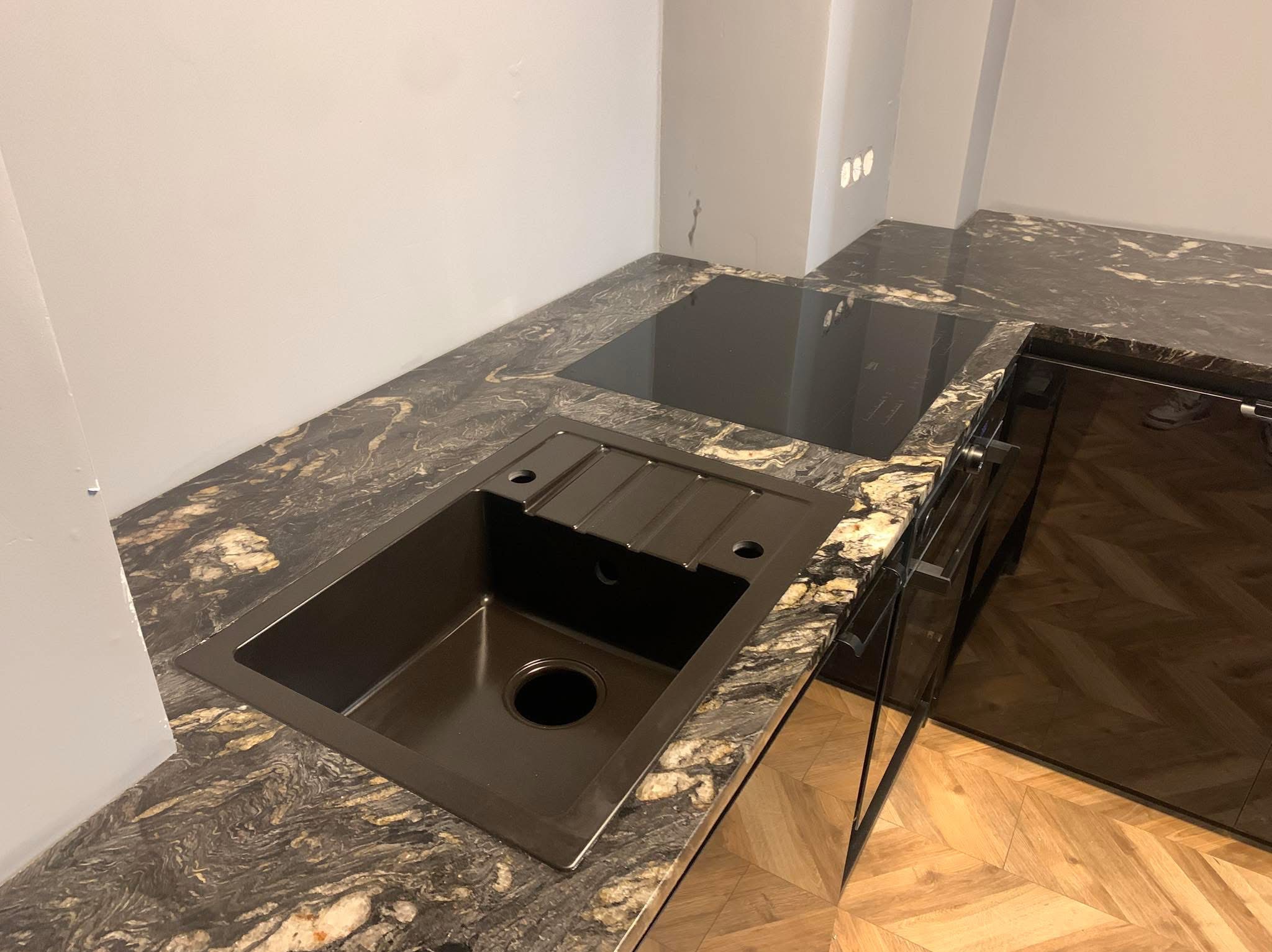 Czarny zlew z ociekaczem wkomponowany w blat kuchenny z ciemnego kamienia z wyraźnym, nieregularnym wzorem, obok płyta indukcyjna i fragment zabudowy kuchennej, widoczna podłoga z parkietu...