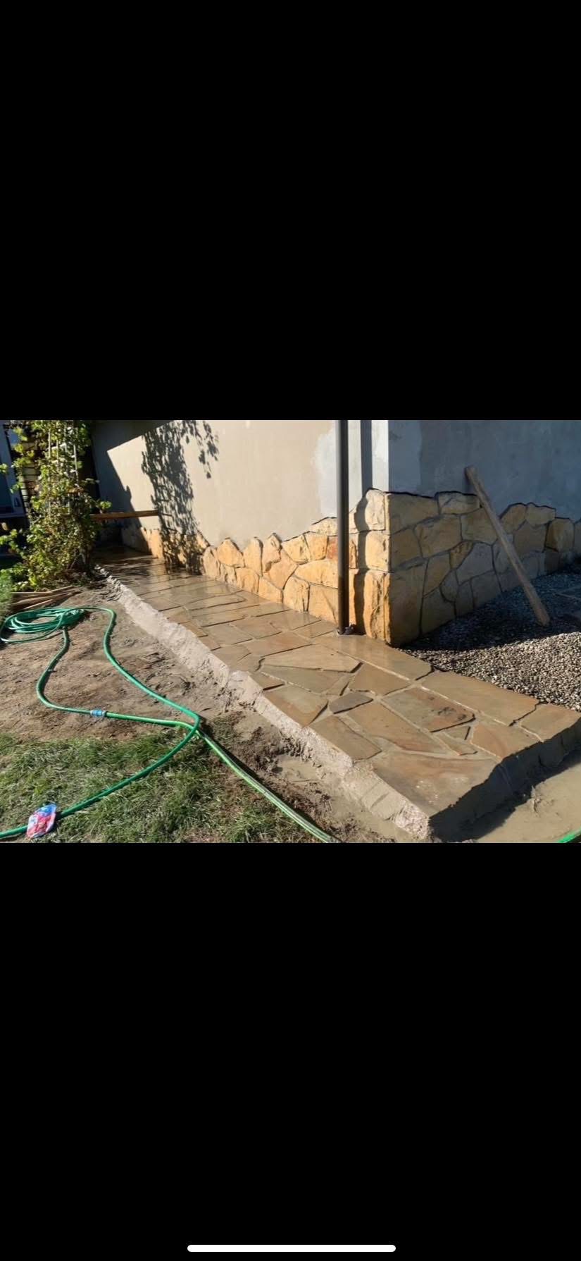 Układanie nieregularnej ścieżki z kamienia naturalnego przy elewacji domu z kamienną podmurówką, widoczny wąż ogrodowy i świeżo zasypany żwir.