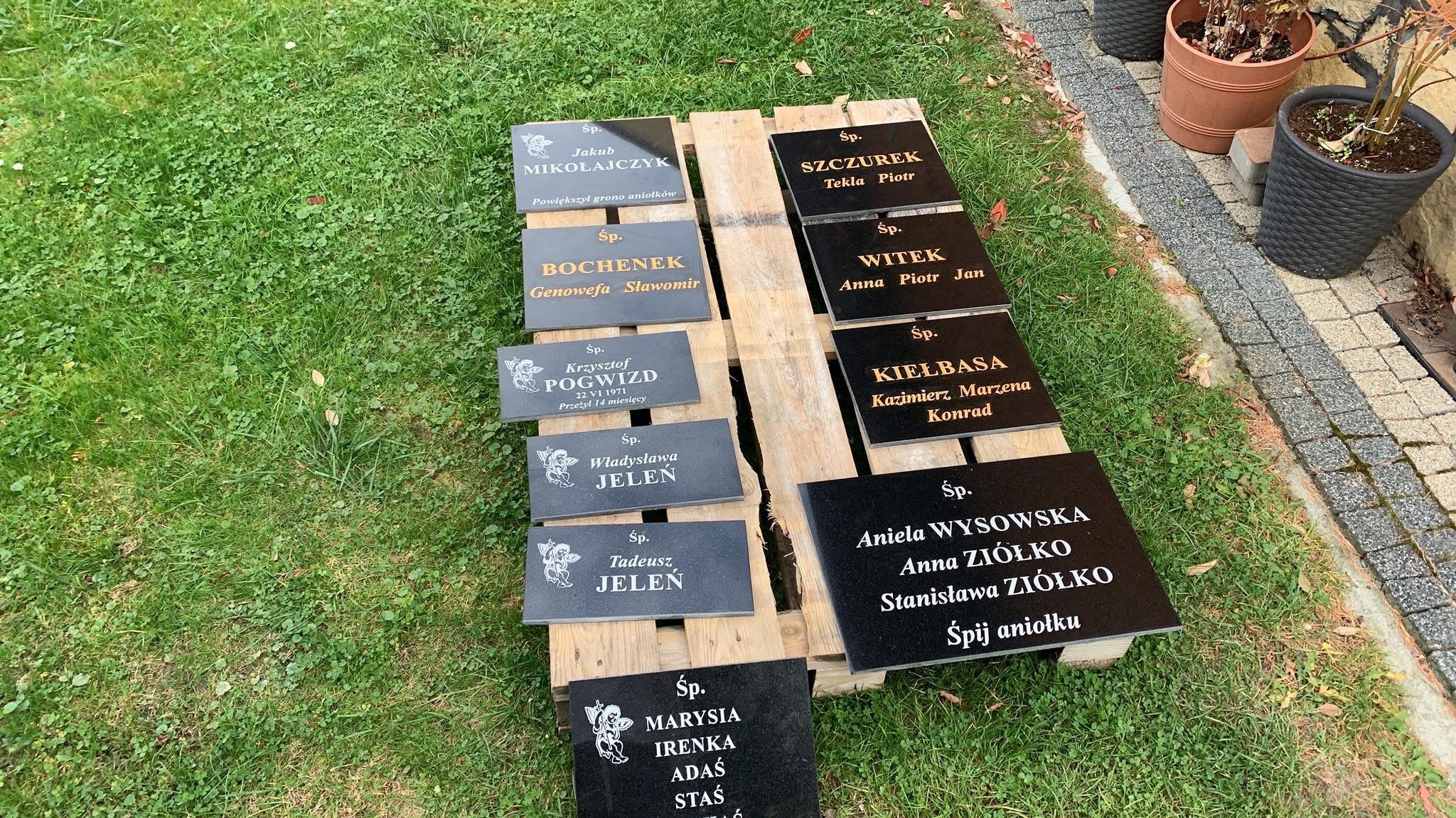 Przygotowane do montażu czarne tabliczki z wygrawerowanymi imionami i nazwiskami, leżące na palecie na tle trawy i kostki brukowej, czekające na umieszczenie na nagrobkach.