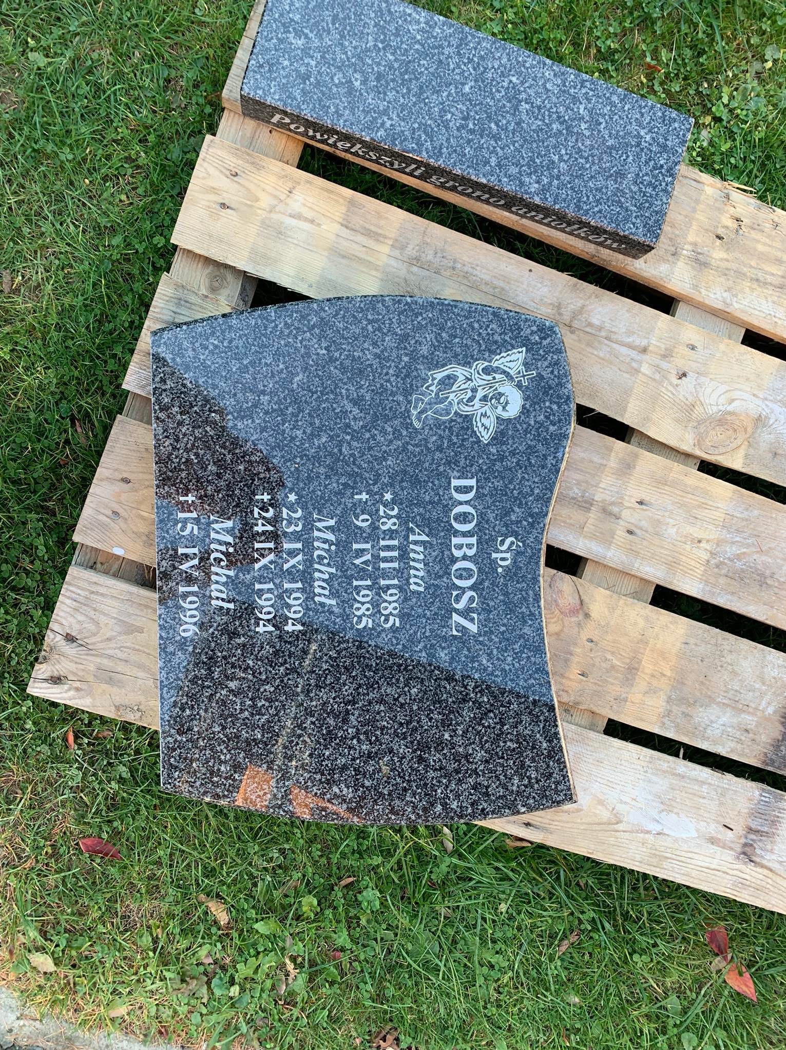 Granitowy element nagrobka z wygrawerowanymi danymi osobowymi i wizerunkiem anioła, leżący na drewnianej palecie na trawie.