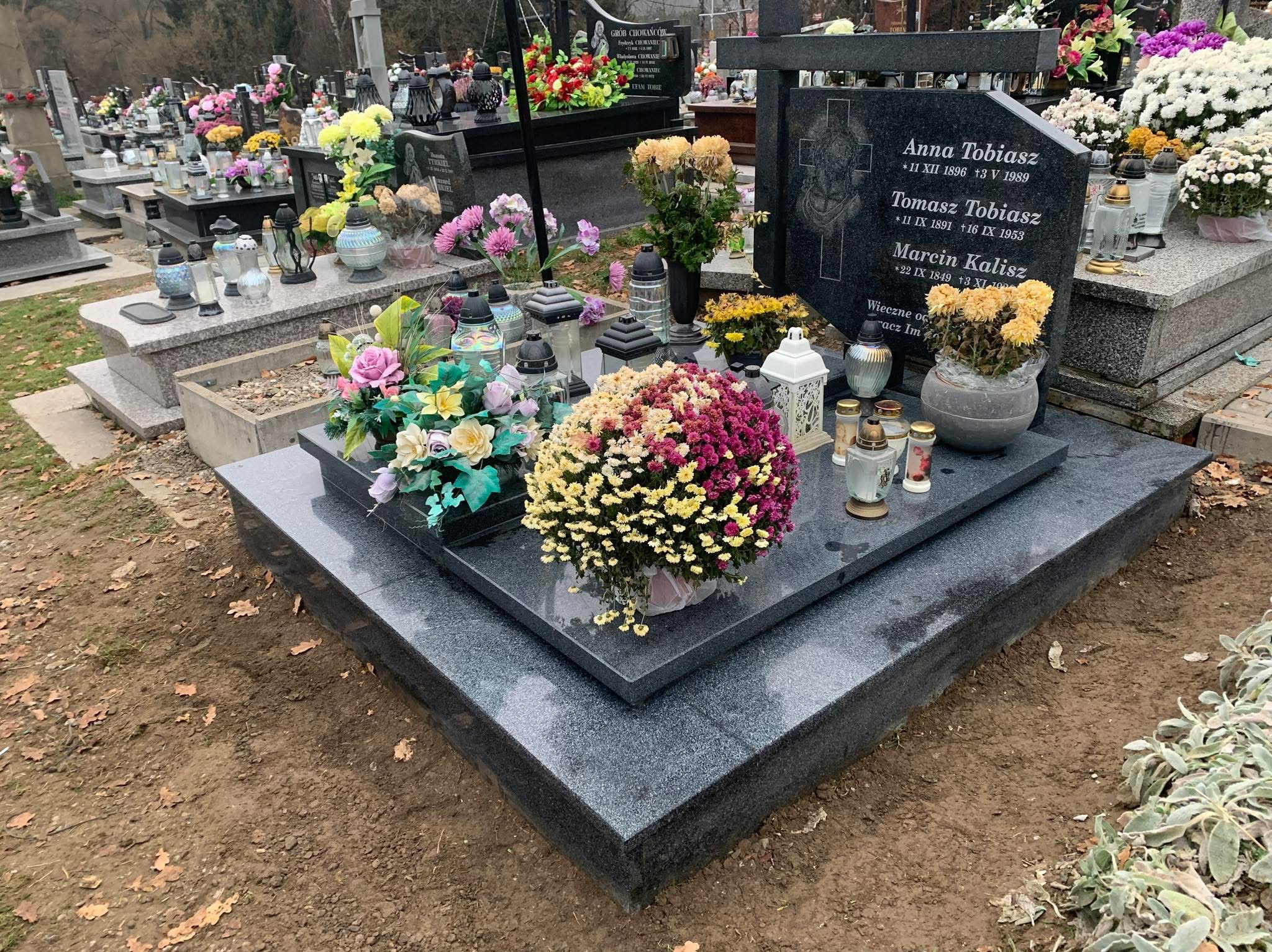 Granitowy nagrobek z inskrypcjami i dekoracjami kwiatowymi oraz lampionami na cmentarzu, widok ogólny na tle innych grobów.