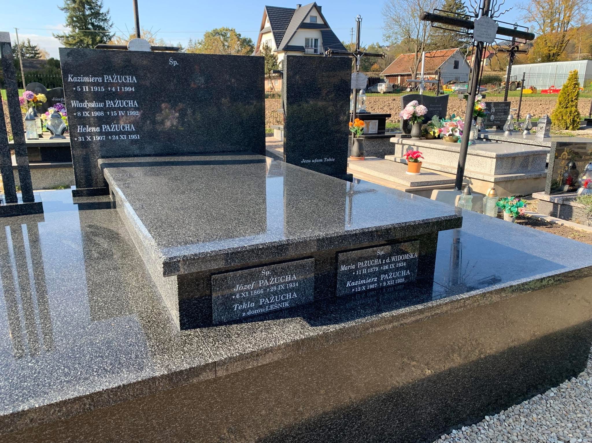 Czarny, polerowany nagrobek z inskrypcjami, widoczny na cmentarzu w słoneczny dzień, z innymi nagrobkami i budynkami w tle.