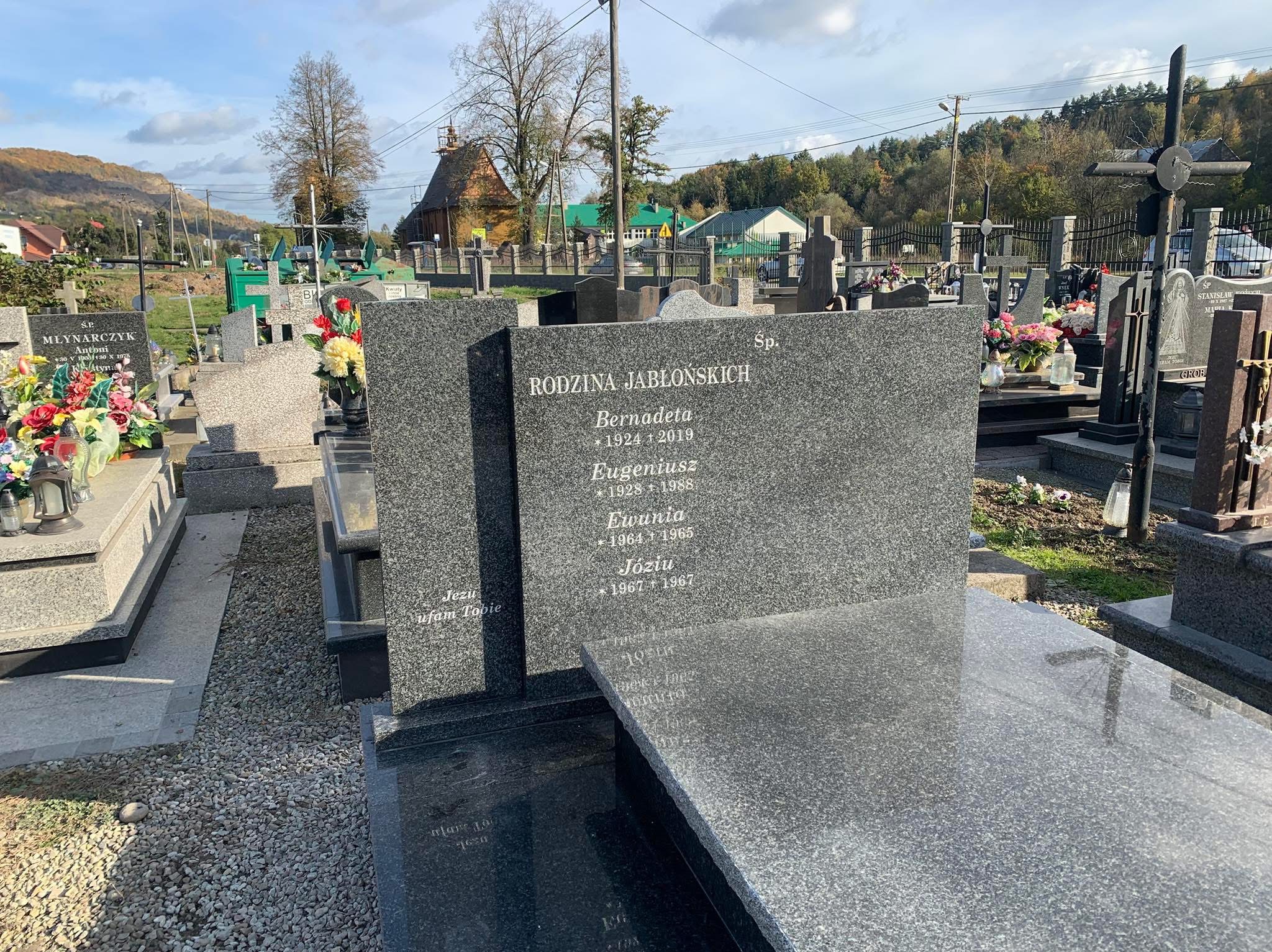 Granitowy nagrobek rodzinny na cmentarzu, z inskrypcjami i datami, otoczony innymi grobami i zielenią, w tle widoczne budynki i drzewa.