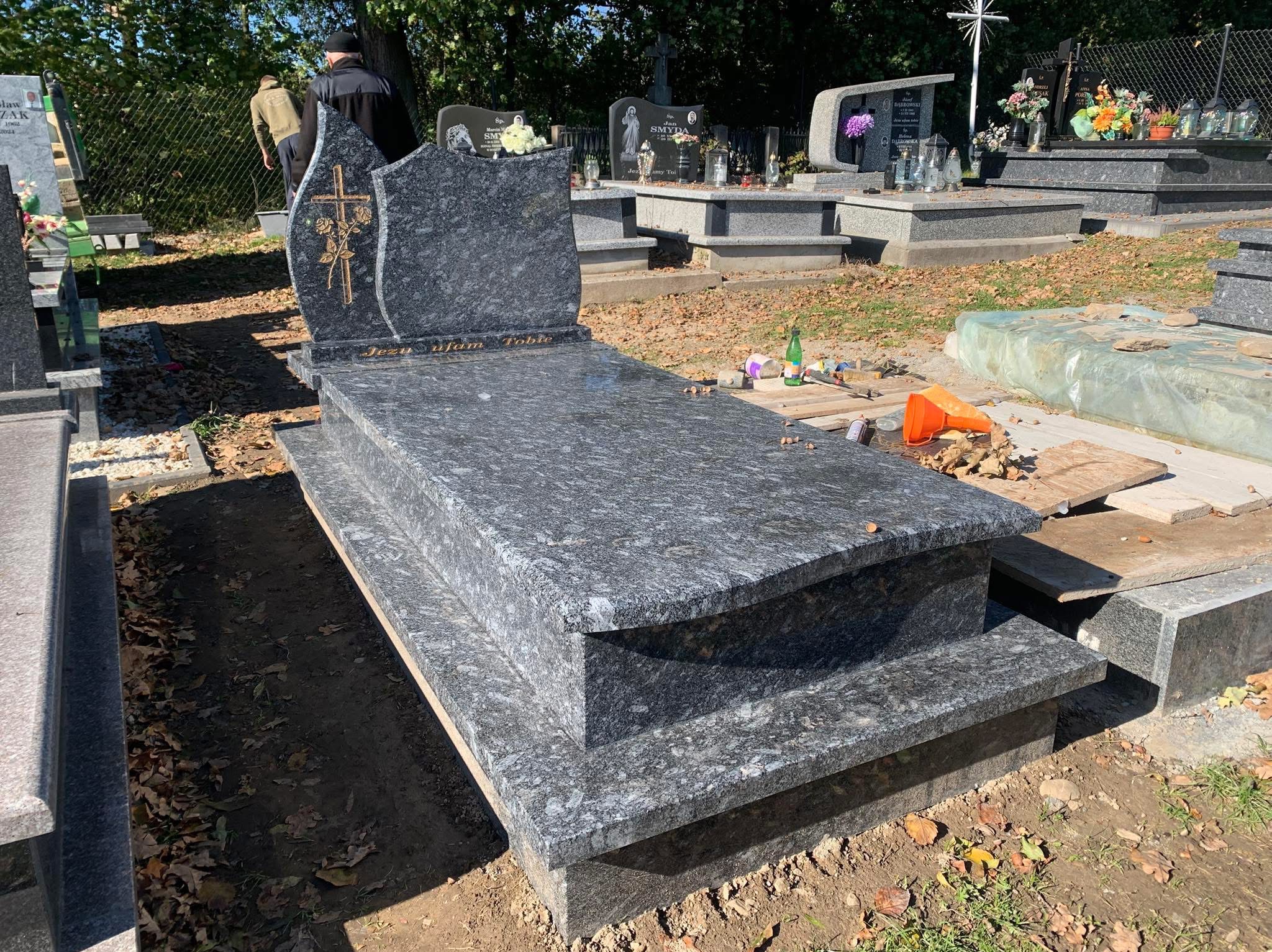 Granitowy pomnik z wygrawerowanym krzyżem i napisem 'Jezu ufam Tobie', otoczony jesiennymi liśćmi na cmentarzu, widoczne narzędzia i materiały budowlane.