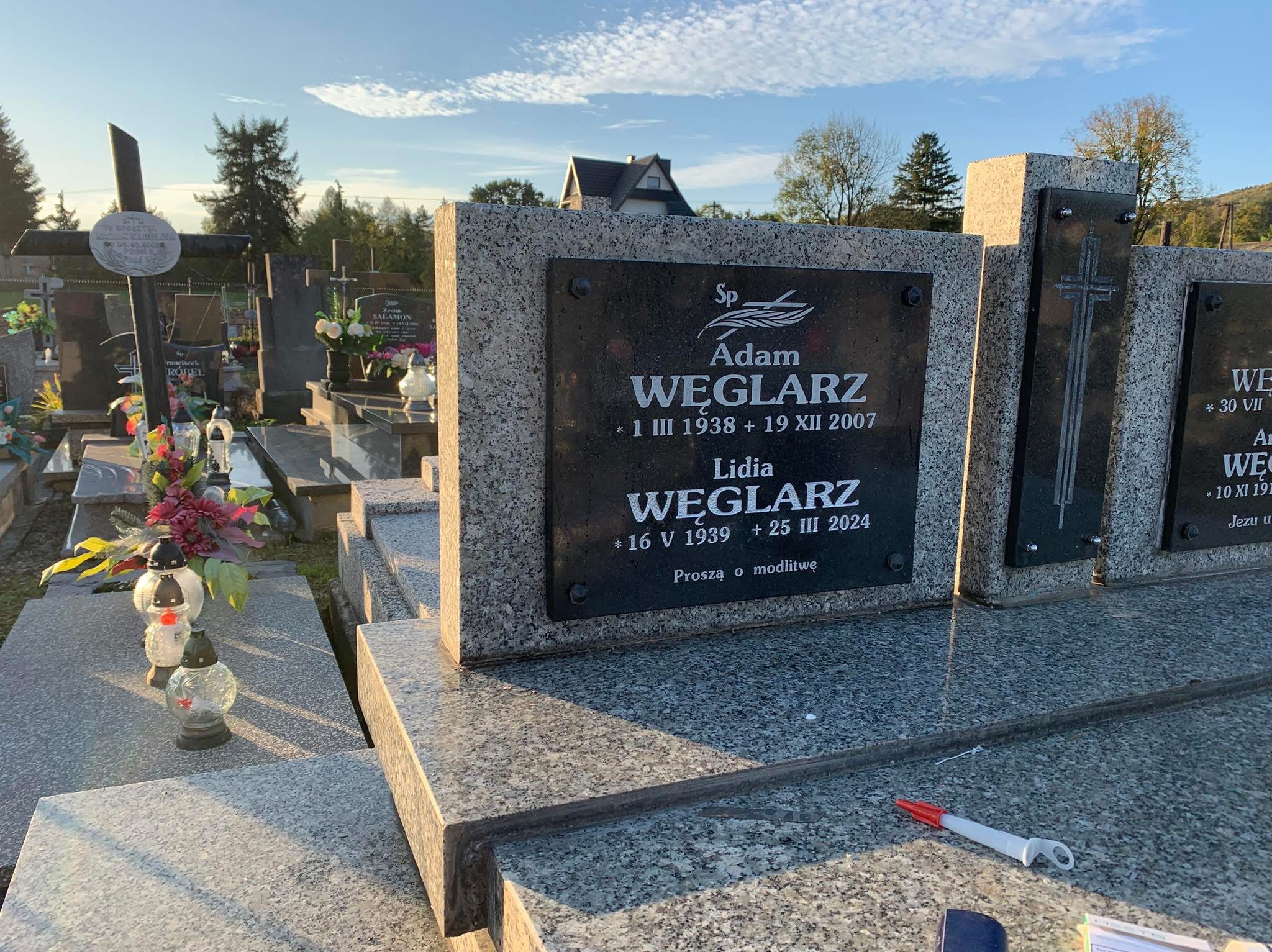 Granitowy nagrobek z czarną tablicą inskrypcyjną z wygrawerowanymi danymi zmarłych, widoczny długopis korekcyjny na płycie nagrobka, cmentarz w tle.