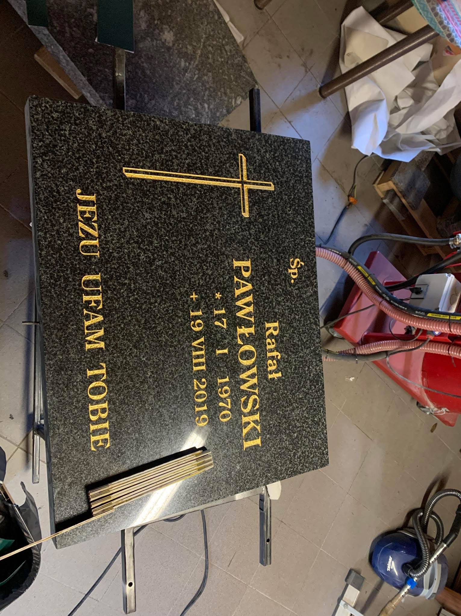 Granitowa tablica nagrobna z inskrypcją w kolorze złotym, zawierająca imię i nazwisko Rafał Pawłowski, daty urodzin i śmierci oraz napis 'Jezu ufam Tobie', ustawiona na metalowym stojaku, w tle...