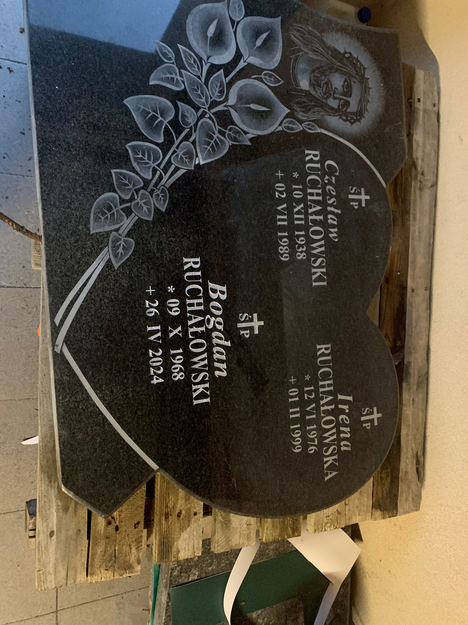 Dwa elementy z ciemnego granitu, prawdopodobnie części składowe pomnika, leżące na drewnianej palecie. Na jednym wyryty wizerunek Jezusa, na obu nazwiska i daty.