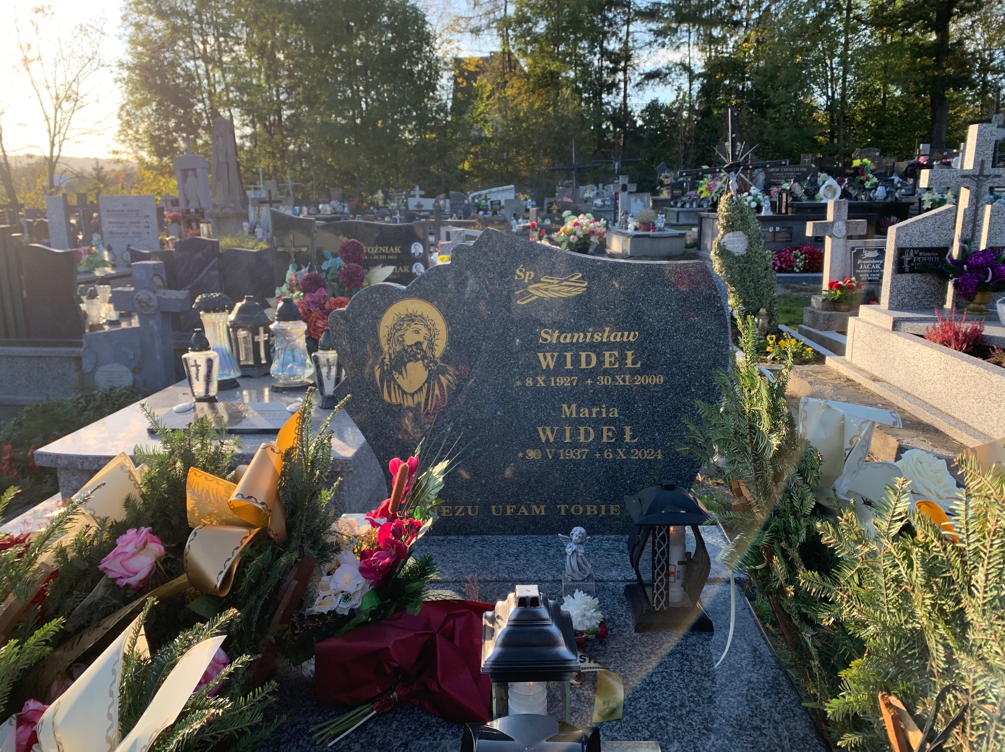 Ciemny, granitowy pomnik z wygrawerowanym wizerunkiem Jezusa i inskrypcjami, ozdobiony kwiatami, wstążkami i lampionami, na tle cmentarza w słoneczny dzień.