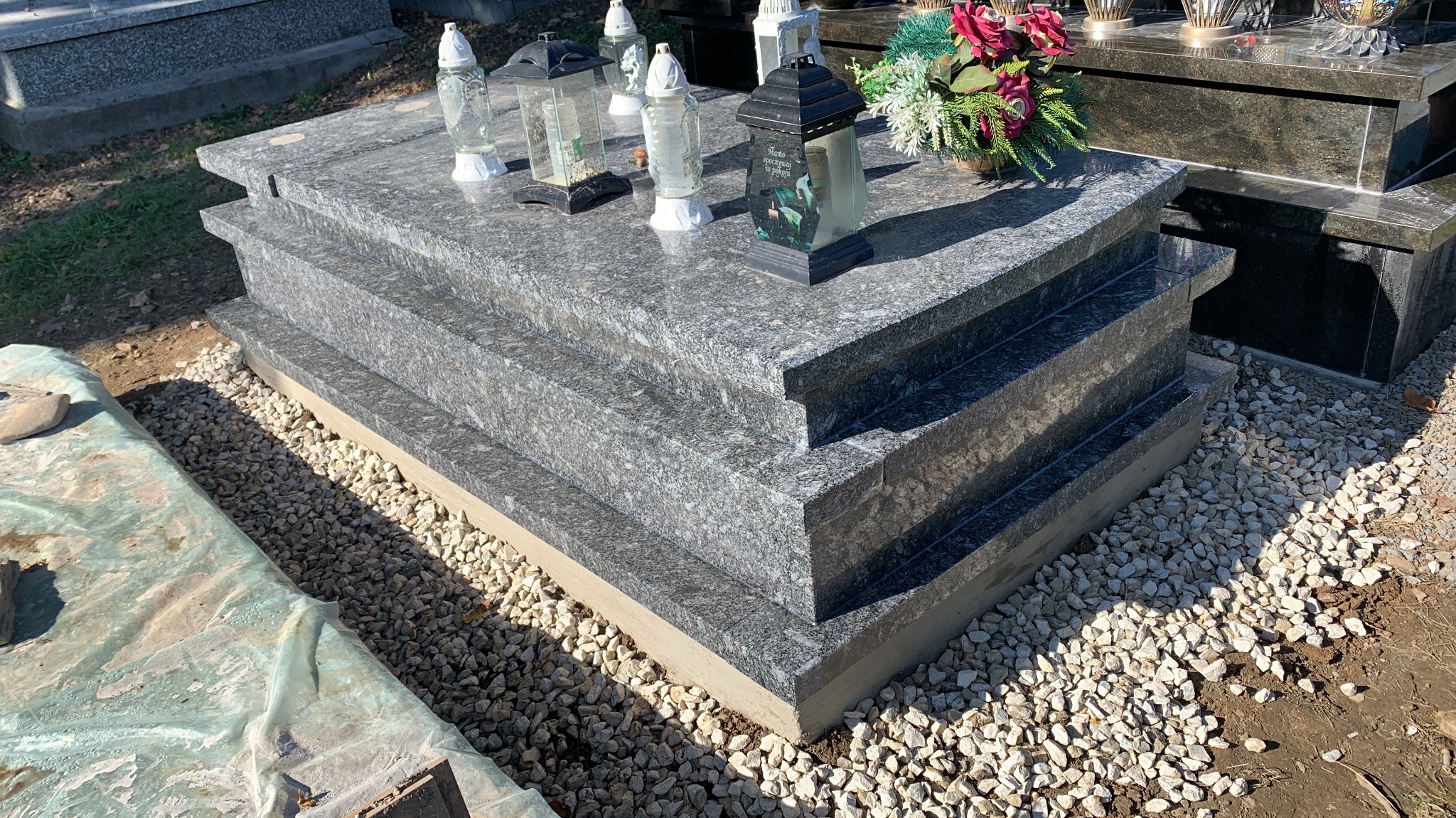 Granitowy pomnik z trzema poziomami, ozdobiony lampionami i kwiatami, otoczony jasnym żwirem.