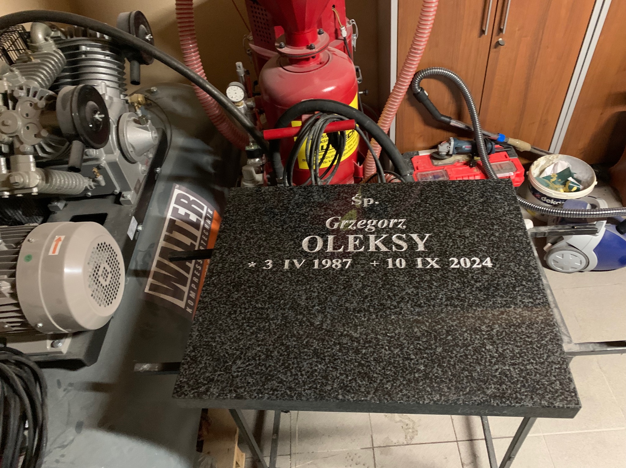 Granitowy nagrobek z wygrawerowanym imieniem i nazwiskiem Grzegorz Oleksy oraz datami urodzenia i śmierci, leżący na metalowym stole warsztatowym w otoczeniu sprężarki, piaskarki i innych narzędzi