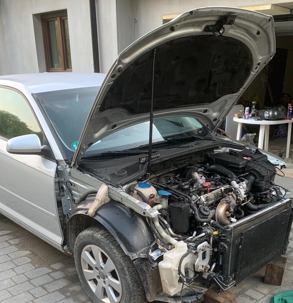 Srebrny samochód z otwartą maską, widoczny silnik w trakcie demontażu elementów, brak przedniego zderzaka i błotnika, na drewnianych klockach pod radiatorem.