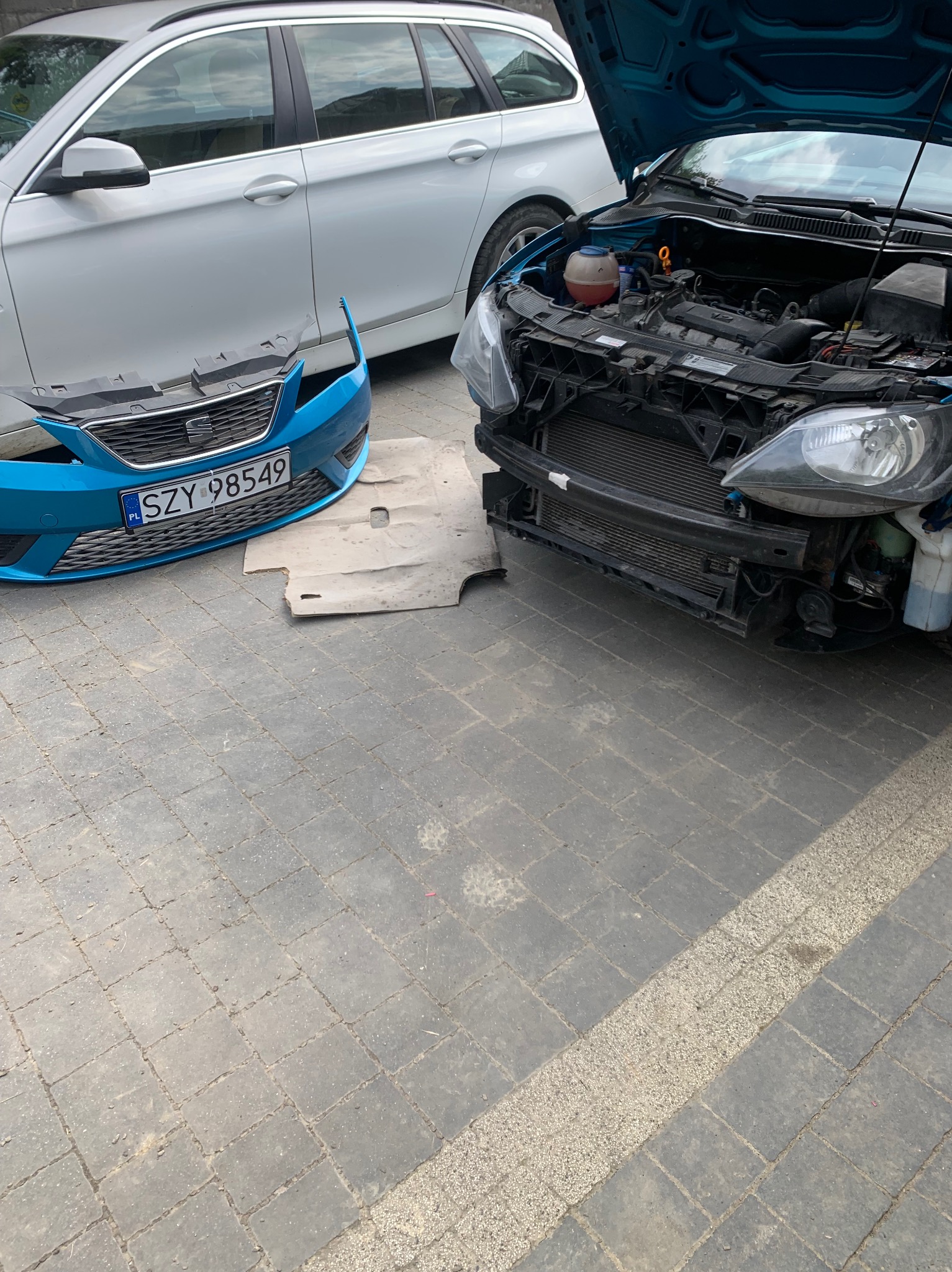 Niebieski samochód z otwartą maską i zdemontowanym przednim zderzakiem, obok białe kombi, widoczne elementy silnika i chłodnicy