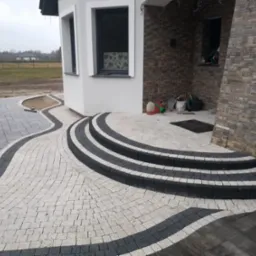 Wykończony podjazd z kostki brukowej w odcieniach szarości i bieli, prowadzący do domu z kamienną elewacją i schodami.