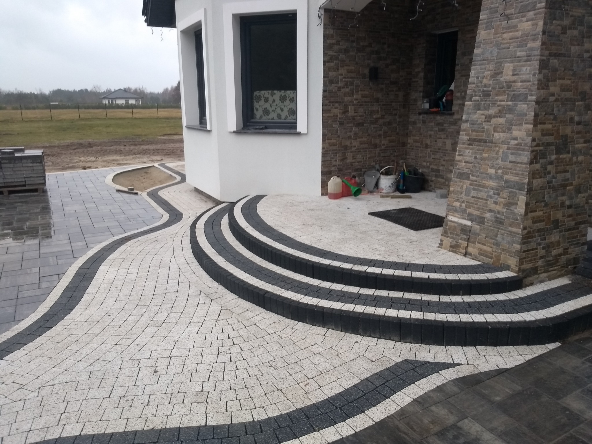 Wykończony podjazd z kostki brukowej w odcieniach szarości i bieli, prowadzący do domu z kamienną elewacją i schodami.