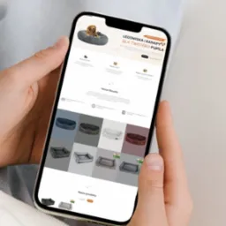 Osoba trzyma telefon prezentujący stronę internetową z ofertą legowisk i kanap dla zwierząt domowych, widoczne różne modele i kolory produktów.
