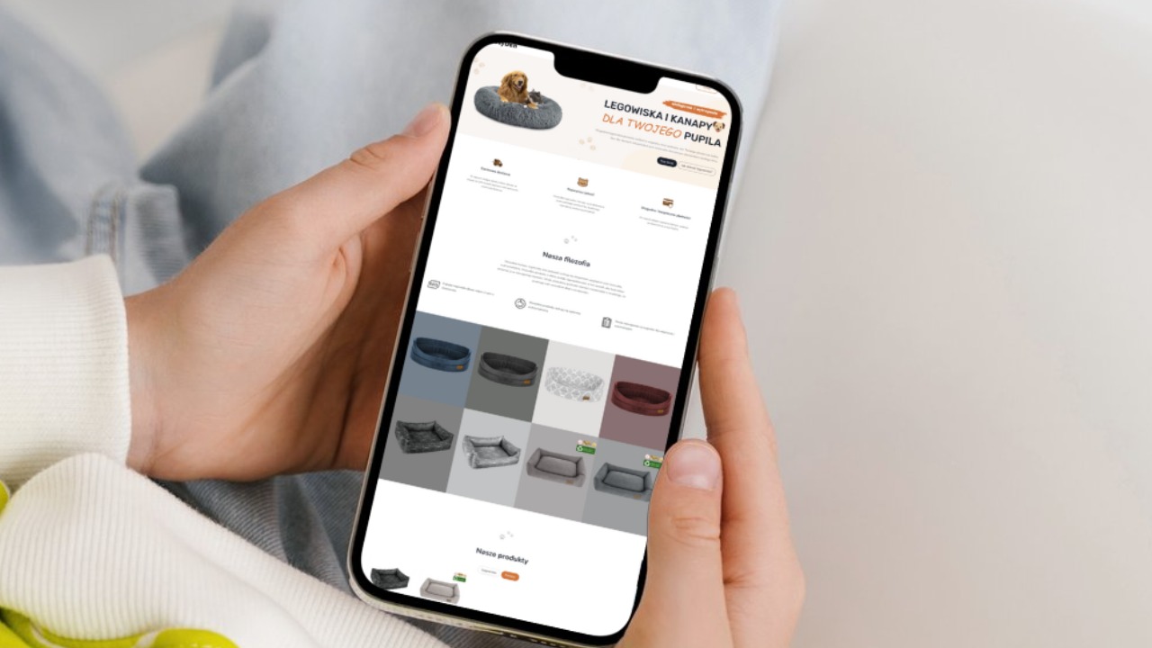 Osoba trzyma telefon prezentujący stronę internetową z ofertą legowisk i kanap dla zwierząt domowych, widoczne różne modele i kolory produktów.