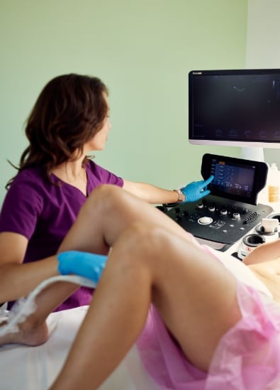 Kobieta w fioletowej bluzie i niebieskich rękawiczkach wykonuje badanie USG nogi pacjentki leżącej na łóżku.