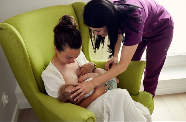 Fizjoterapeutka w fioletowym uniformie pomaga młodej matce karmiącej piersią niemowlę w jasnozielonym fotelu, drugie dziecko siedzi obok.