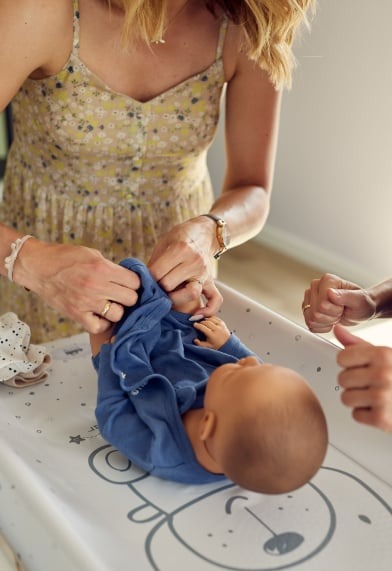 Osoba w kwiecistej sukience ubiera manekina niemowlaka w niebieskie ubranko na przewijaku z motywem misia.