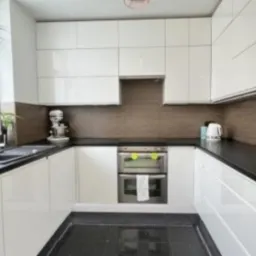 Biała, nowoczesna kuchnia w kształcie litery U z czarnymi blatami i podłogą z połyskiem, widok na okno z wiszącą rośliną i sprzęt AGD.