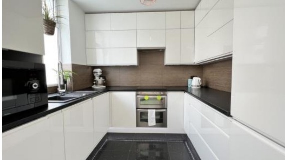 Biała, nowoczesna kuchnia w kształcie litery U z czarnymi blatami i podłogą z połyskiem, widok na okno z wiszącą rośliną i sprzęt AGD.