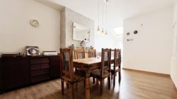 Jasna jadalnia z drewnianym stołem i krzesłami, parkiet w odcieniach brązu, komoda z dekoracjami i zegar na ścianie, oświetlenie wiszące nad stołem.