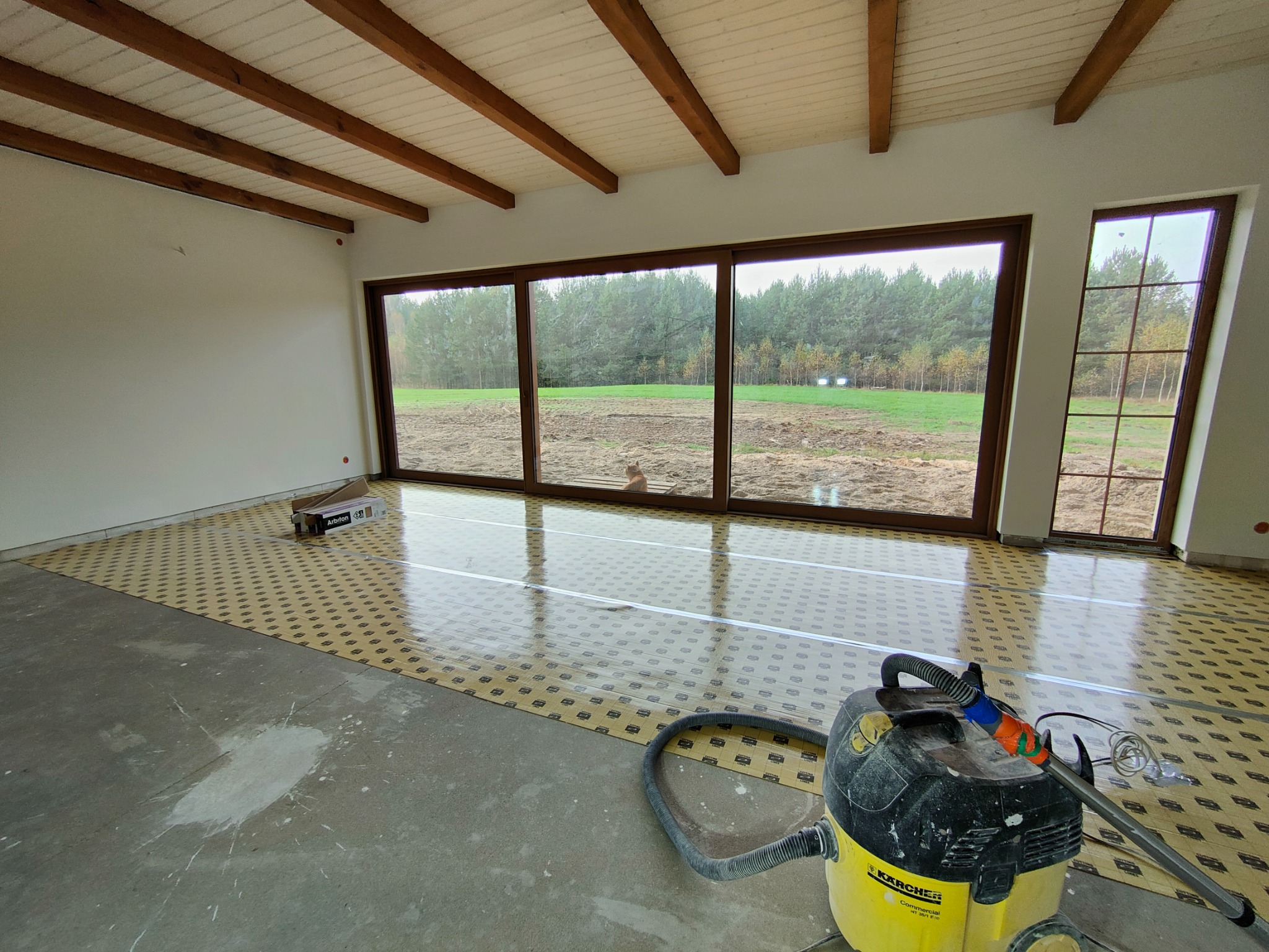 Przygotowanie podłoża pod panele: rozłożona folia izolacyjna z widocznym opakowaniem produktu Arbinton, odkurzacz przemysłowy Karcher w narożniku, surowy beton i białe plamy na podłodze, duże okna...